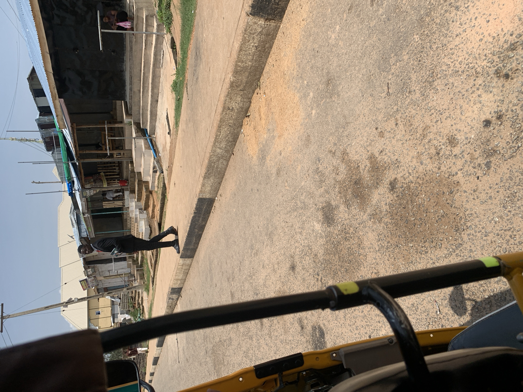In Gura Top, Nigeria, a young man in a black suit and tie walks on the sidewalk in front of a row of small shops. The shops have corrugated metal roofs and open fronts. To the right, a woman sits on steps outside a shop holding a pink bag. The ground is paved with asphalt and a concrete sidewalk. A yellow auto-rickshaw is partially visible in the foreground. The sky is clear and blue.