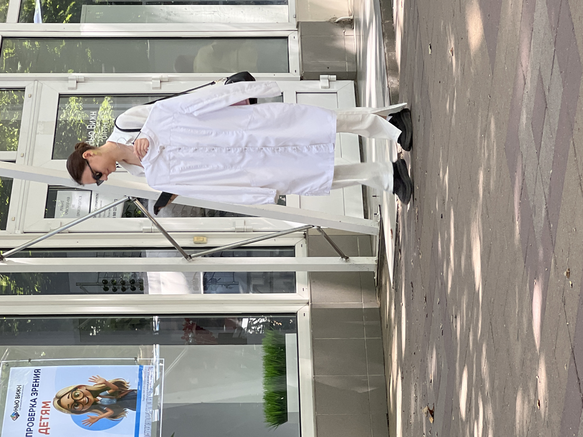 A woman stands outside an optical shop in Krasnodar, Russia, holding a white lab coat. She is wearing white pants and black shoes. The shop has a glass front with posters displayed, one of which features an illustration of a cartoon girl wearing glasses. The ground is paved with grey tiles, and sunlight casts dappled shadows. The overall impression is that of a pedestrian walkway adjacent to a commercial establishment on a sunny day.