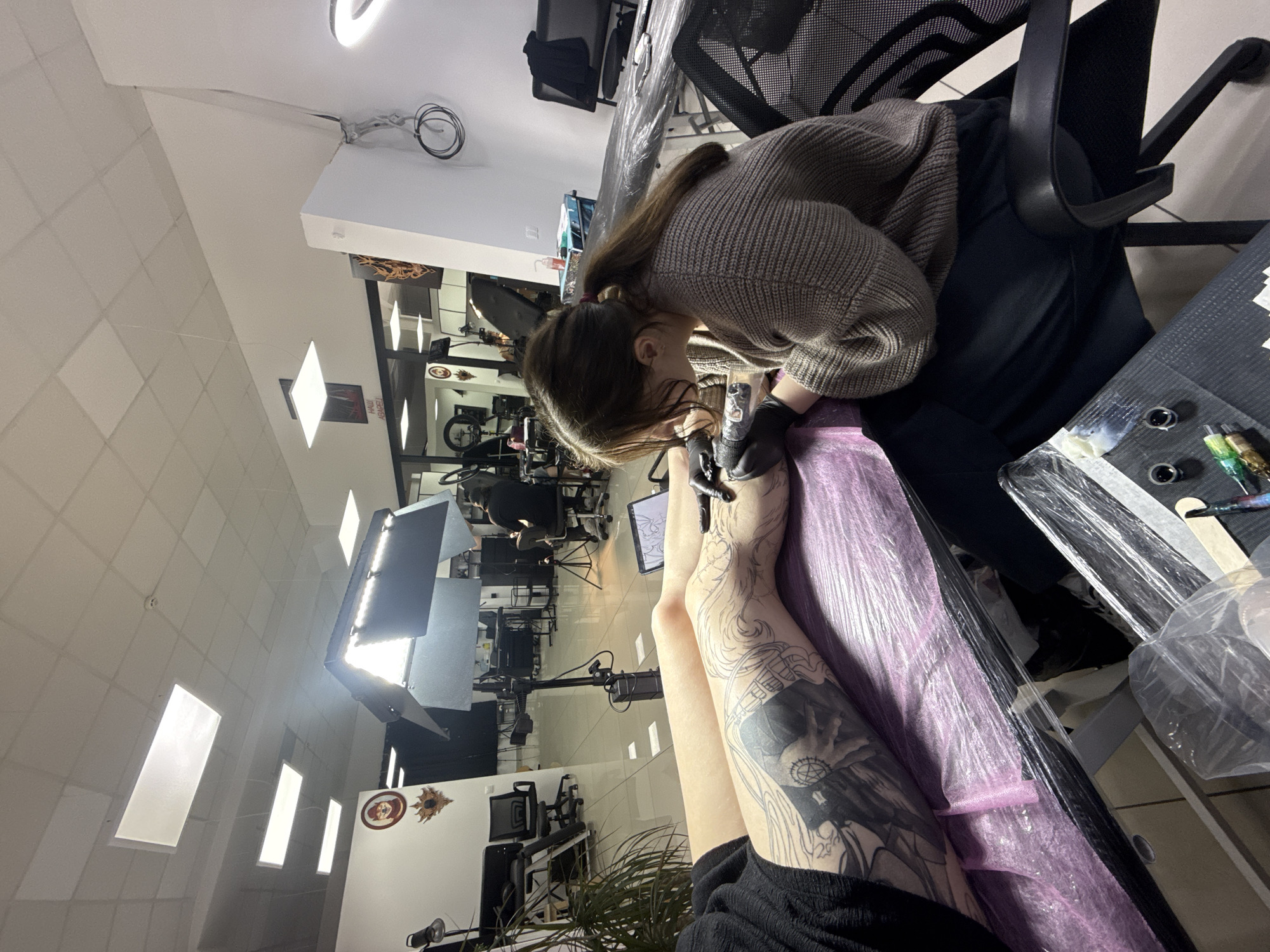 The image shows a woman giving a tattoo to another person in a tattoo parlor in Unknown, Unknown. The tattoo artist is wearing a sweater and black gloves and is focused on her work. The person receiving the tattoo has several tattoos on their legs. The tattoo parlor appears to be clean and well-lit. A table with ink and other supplies is next to the tattoo recipient.