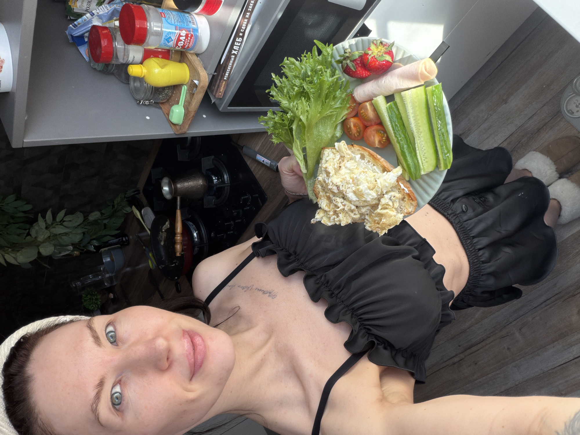 A woman is taking a selfie while holding a plate of food. The plate contains lettuce, strawberries, ham, sliced tomatoes, sliced cucumbers, and a sandwich with a white filling. The woman is wearing a black ruffled top and black pants. She has fair skin and light-colored eyes.
Behind her, there is a kitchen counter with various food items, including spices and a bottle of mustard. There is also a microwave and a stove with a cezve.
Based on the presence of the product "ХЛЕБ И СДОБА НА ЗАКВ" which translates from Russian to "BREAD AND BUNS ON STARTER", referring to a product made by В. Войнова, and referencing that it's sold at a store located in Perm, Russia, the photo was taken in Perm, Russia.