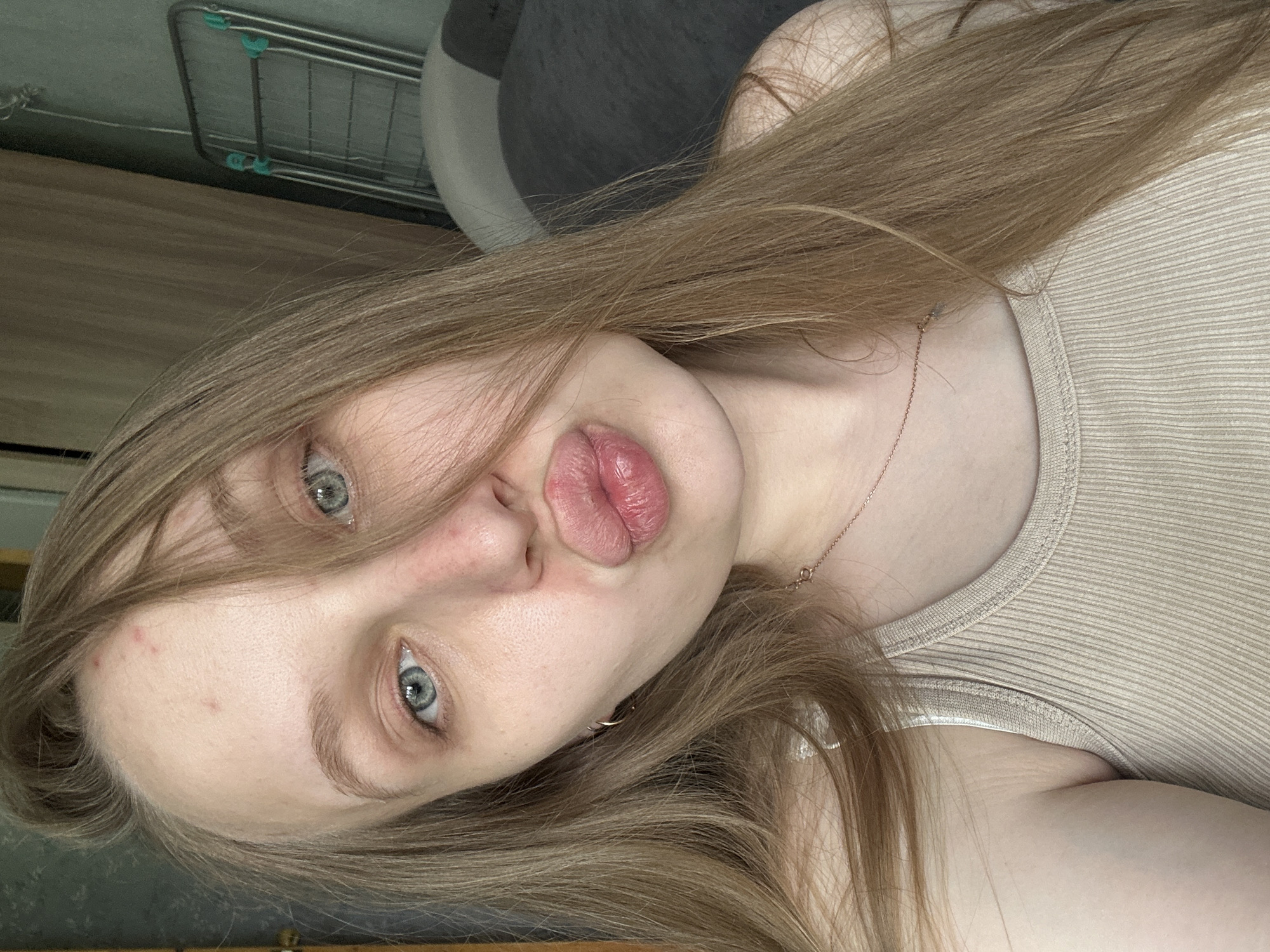 A young woman with long, light brown hair is pictured indoors. She is wearing a light-colored ribbed tank top. She has light skin and light blue eyes. She is facing the camera with pursed lips. A thin gold necklace is visible around her neck. In the blurred background, there is a clothing rack and some dark furniture. The image does not provide enough information to determine the location in the city of Chaikovsky, Russia.