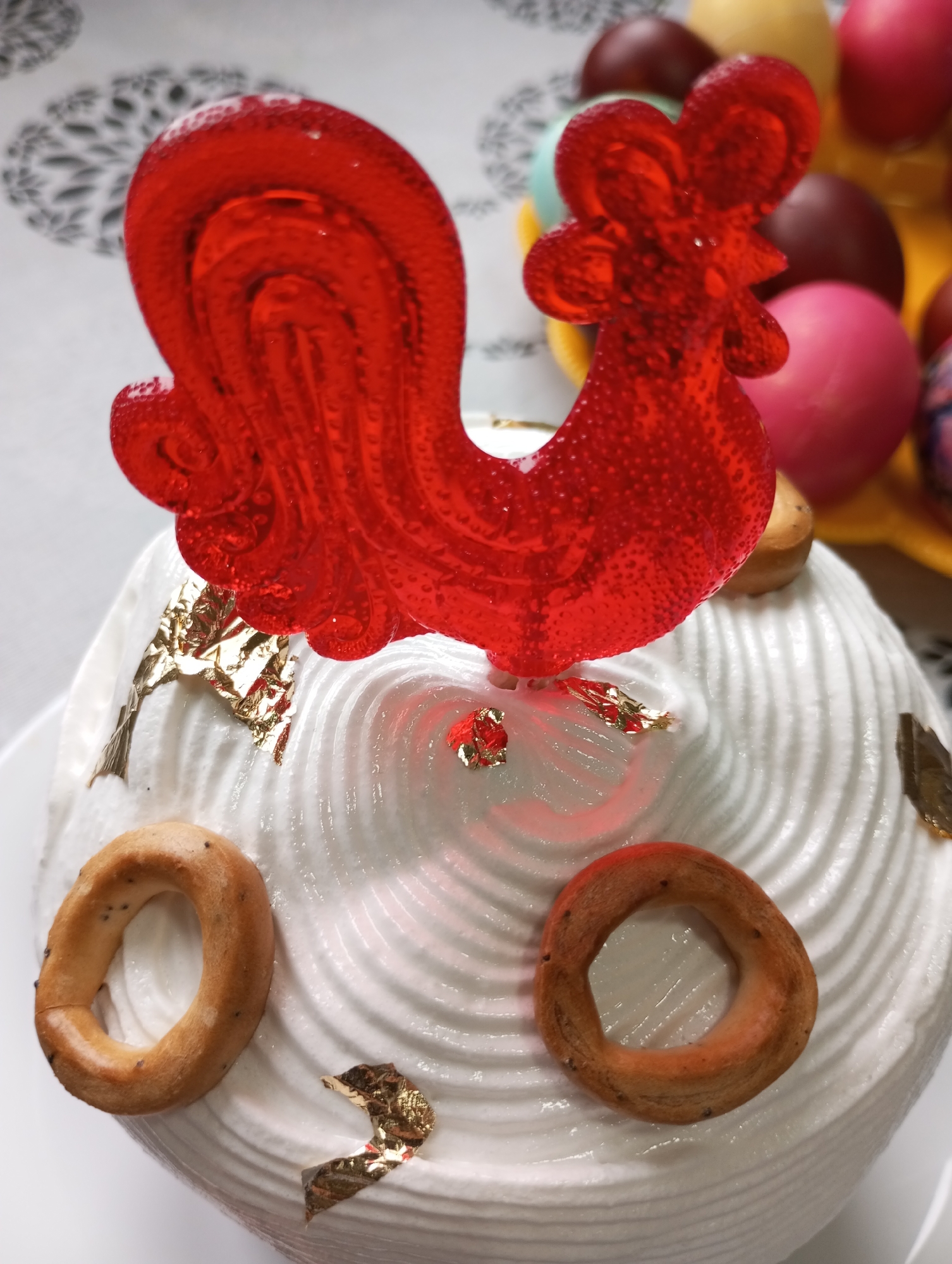 A close-up shot shows a decorated cake with a red, rooster-shaped lollipop on top. The cake is covered in white, swirled icing and adorned with two ring-shaped pastries, and gold leaf pieces. In the background, there is a collection of colored eggs. The image was taken in Буренка, Russia.