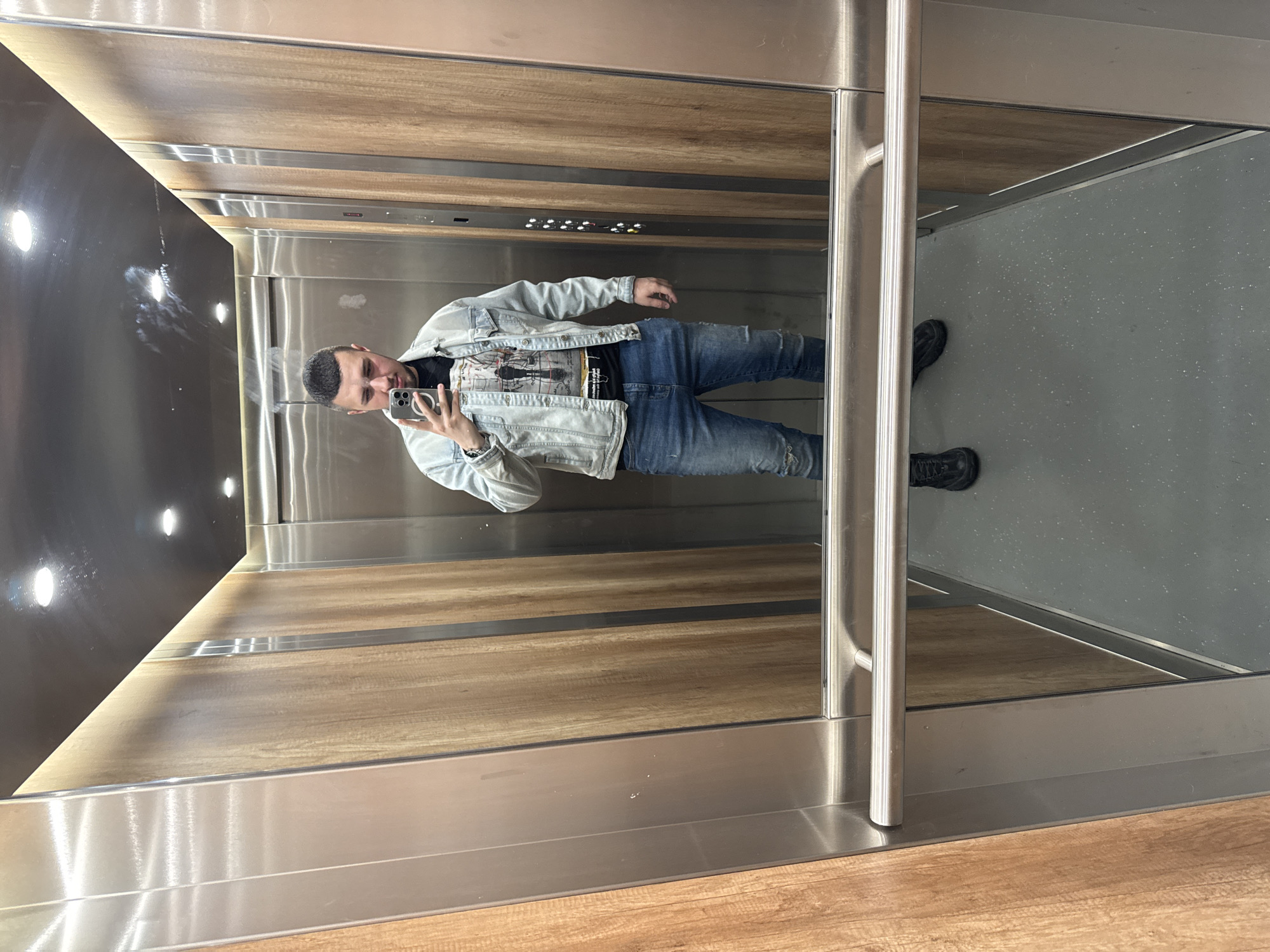 A man is captured in a mirror selfie inside a modern elevator, appearing to be in Sofia, Bulgaria. He stands facing the mirror, holding a smartphone in his right hand to take the picture. He has short, dark hair and a neatly trimmed beard, with a neutral to slightly smiling expression.
He is dressed in casual attire: a light blue denim jacket layered over a black t-shirt featuring a detailed graphic design that includes what appears to be an urban landscape or abstract art, with faint, unreadable text or signatures beneath it. He wears blue ripped jeans and black athletic or hiking-style shoes. A watch is visible on his left wrist.
The elevator's interior is clean and contemporary. One wall, where the selfie is being taken, is entirely mirrored, reflecting the man and the opposite wall. The walls behind the man are paneled with light brown wood-look material, accented by horizontal brushed stainless steel strips. A control panel with several illuminated buttons is integrated into the wooden section. The ceiling is dark, possibly black, with multiple recessed spotlights, some of which create bright reflections on the mirrored surface. A horizontal brushed metal grab bar runs across the mirror wall. The floor of the elevator is a light grey speckled material. The image itself is oriented horizontally, depicting the man standing upright within the mirrored reflection.