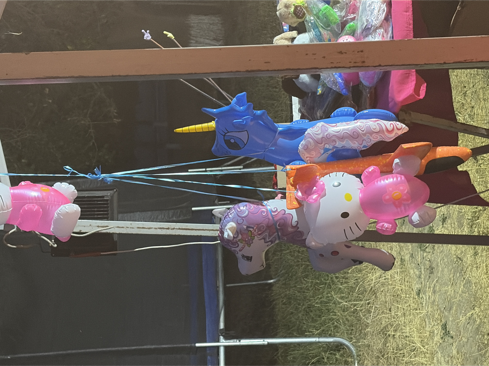 The image shows a collection of inflatable toys, including a Hello Kitty balloon, a unicorn balloon, and other animal-shaped balloons, suspended by blue strings. The toys are illuminated by artificial light, suggesting it is nighttime. In the background, a trampoline and a table laden with more toys can be seen. A portion of a wooden post and a fence are also visible. The scene appears to be set up for a fair or event. No people are visible in the image.