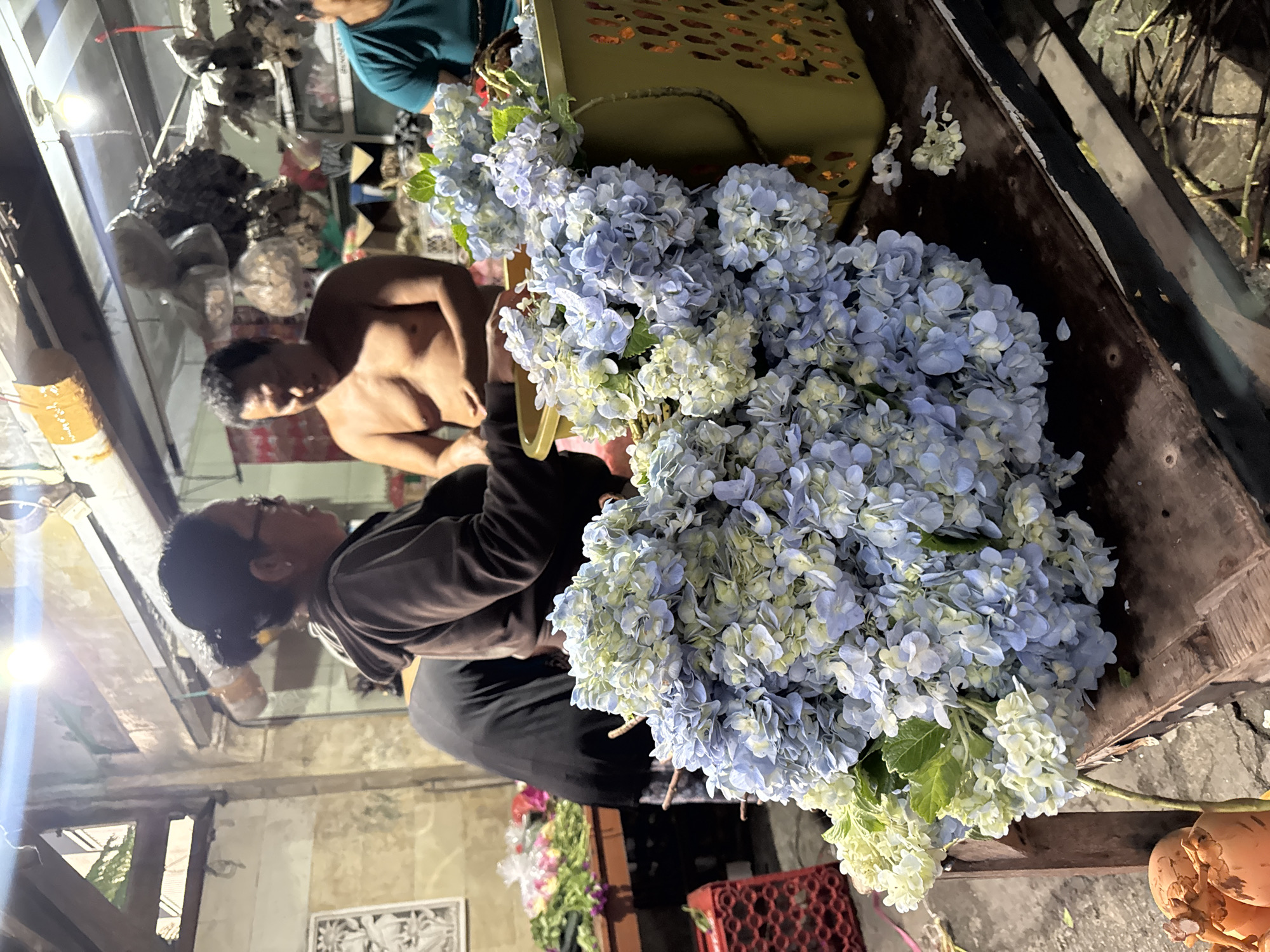 The image shows a flower stand in Kuta, Indonesia. A large bouquet of blue and white hydrangeas dominates the foreground. A man in a brown hooded jacket appears to be working at the stand. Behind him, a shirtless man is also present. Other people are partially visible in the background. A green basket is positioned next to the hydrangeas. The setting seems to be a street with an aged building.