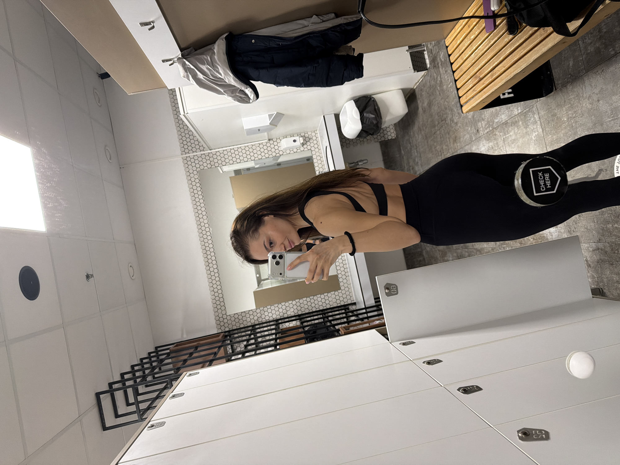 The image depicts a woman taking a selfie in what appears to be a locker room. She is wearing a black sports bra and leggings and is holding a cell phone in her left hand. In her right hand, she is holding a black weight. The background includes rows of white lockers, a mirror, and bathroom facilities. The location is not specified in the image but appears to be in a gym or fitness center. The city of Varna, Bulgaria is not specified in the image.