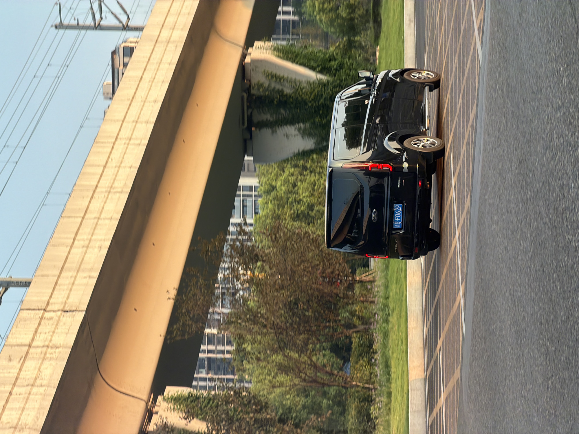 The image shows a black Ford Transit van driving on a road in Jiangjin District, Chongqing, China. The van is traveling away from the camera, and its license plate is visible. In the background, there is an elevated concrete structure, possibly a bridge or overpass, with power lines running overhead. Behind the elevated structure, there are buildings and trees, suggesting an urban or suburban environment. The sun is setting, casting a warm glow on the scene. The road surface has lane markings.