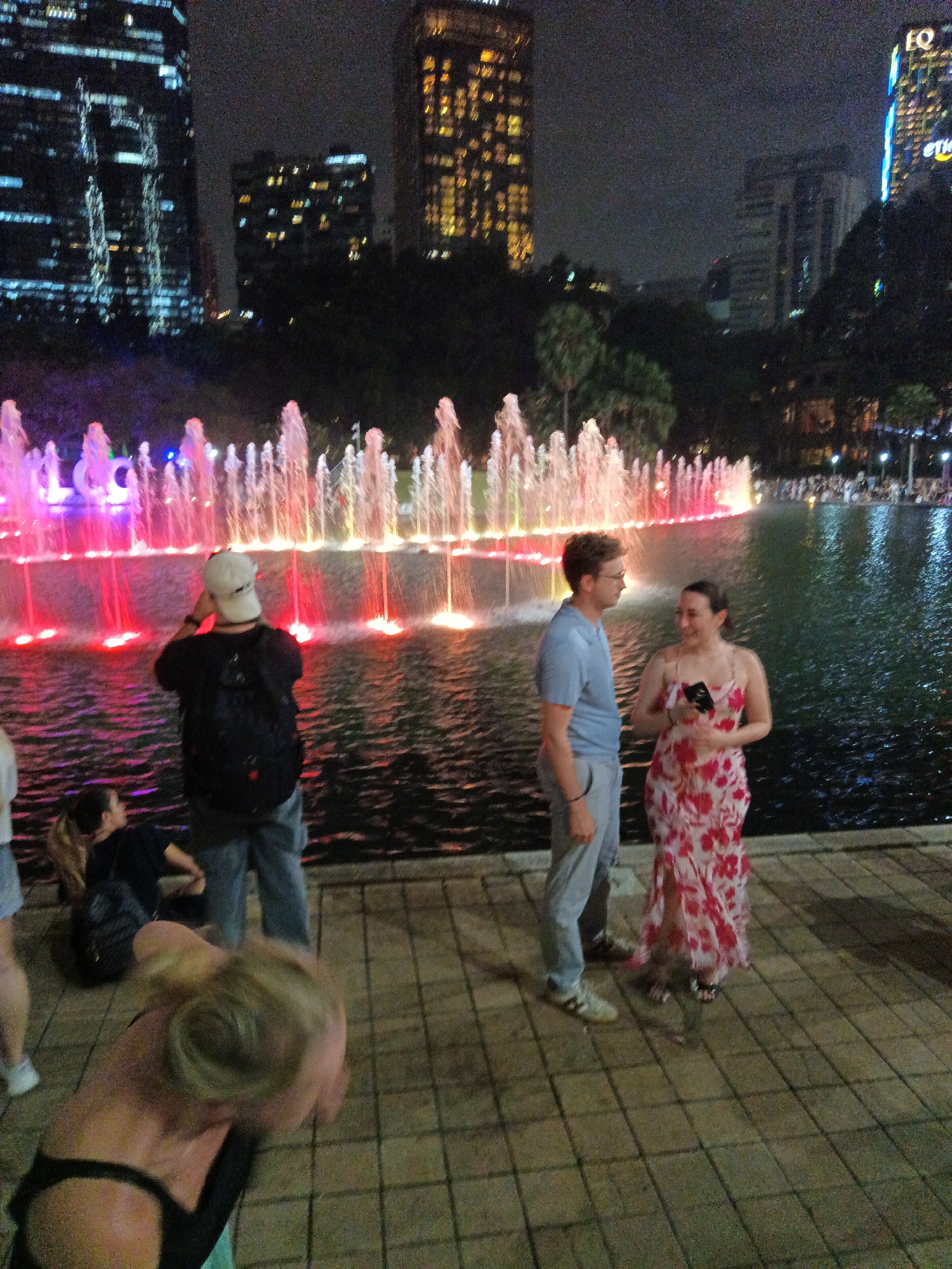 This photograph captures a nighttime scene at a fountain area in Kuala Lumpur, Malaysia. The background features illuminated skyscrapers, a hallmark of the city's skyline, with lights from buildings casting a glow. In the foreground, a group of people are gathered near a large, brightly lit fountain. The fountain is characterized by numerous streams of water illuminated with red and pink lights, creating a vibrant spectacle.
To the left, a man wearing a black t-shirt, grey shorts, and a white cap is seen from the back, facing the fountain. He appears to be wearing a backpack. Beside him, a woman with long brown hair, wearing a dark top, is partially visible.
In the center of the image, a man in a light blue t-shirt and grey pants is standing and looking towards his right. Next to him, a woman in a flowing red and white floral dress is engaged in conversation, holding a dark object in her hand.
Further to the left and slightly out of focus, a person's blonde hair is prominently visible in the foreground, suggesting they are leaning or sitting. The ground is paved with square tiles. The overall atmosphere is festive and dynamic, typical of a public space at night with a water feature.