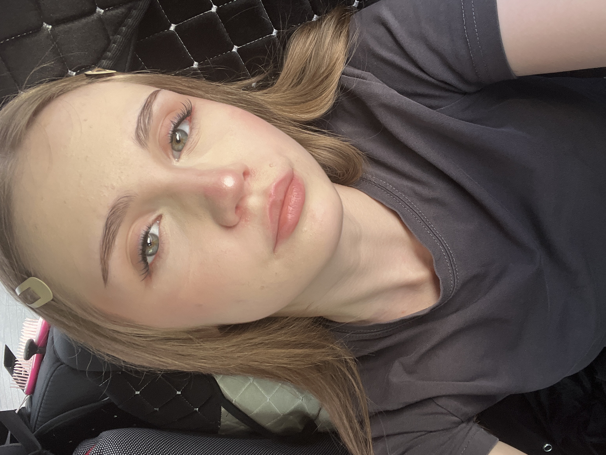 A young Caucasian woman with fair skin, light brown hair, and green eyes is shown from the chest up. She is wearing a dark grey t-shirt. Her hair is parted in the middle and falls around her shoulders. She has a light makeup application, with blush on her cheeks and nose, and mascara on her eyelashes. Two light brown hair clips are visible in her hair. She is looking directly at the camera. The background appears to be the interior of a car, with a dark, quilted seat visible behind her and a glimpse of what looks like a car seat or headrest on the left. The lighting suggests an indoor setting, possibly during the day, with soft light illuminating her face. There is no discernible location in the city of Загородный, Russia.