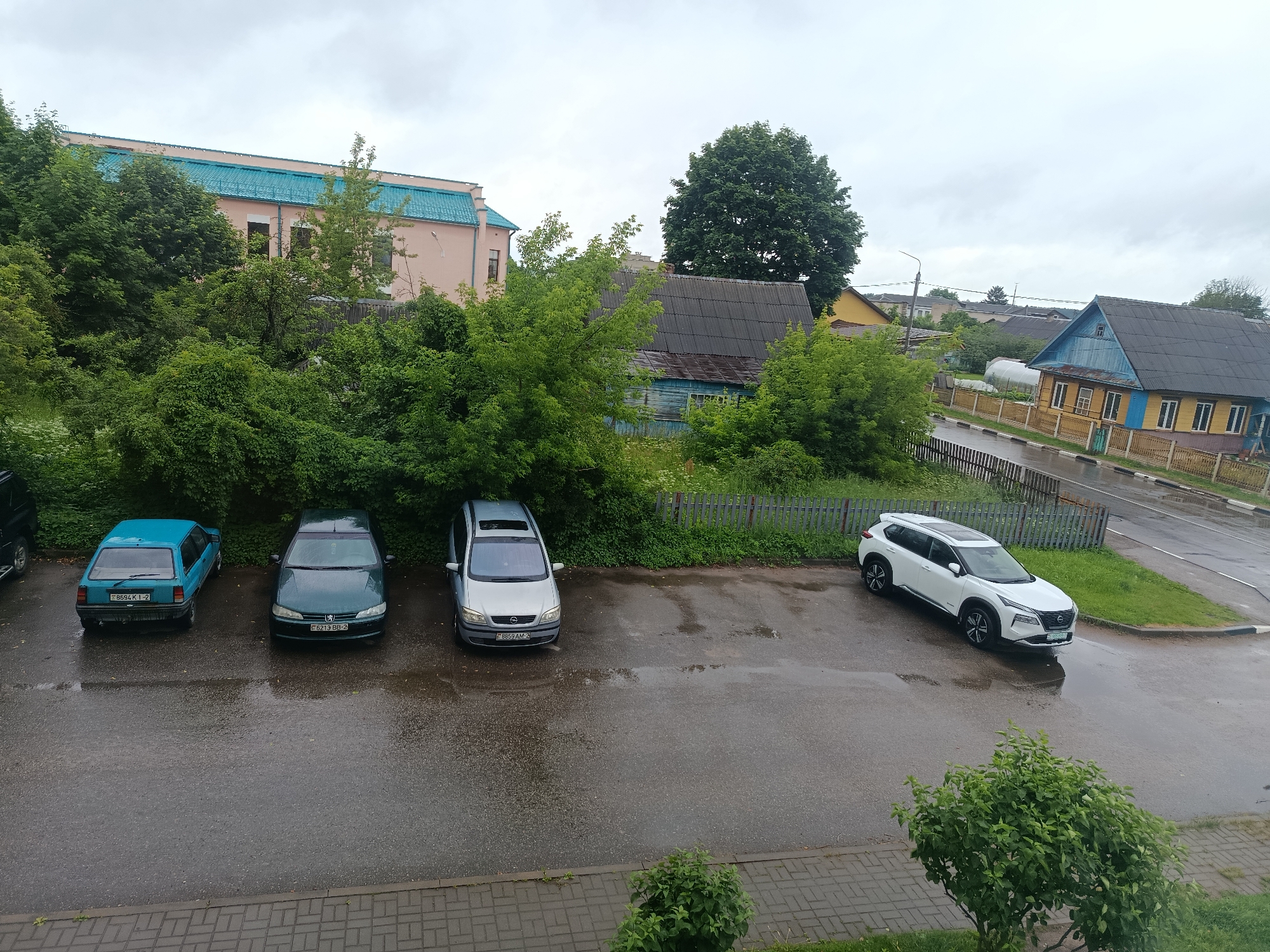 The image depicts an overcast day in Lyepyel, Belarus, with a view of parked cars on a wet asphalt surface adjacent to a sidewalk. In the foreground, a paved sidewalk with a brick pattern is visible, bordered by green bushes. A parking lot extends from the sidewalk, occupied by four cars: a blue hatchback, a dark green sedan, a silver minivan, and a white SUV. The wet asphalt indicates recent rain. Behind the parking lot, a wooden fence separates the area from a grassy strip and dense green trees. Further back, various buildings are visible, including a light pink building with a teal roof, a blue wooden house, and a yellow house with a dark roof. The sky is cloudy and grey.