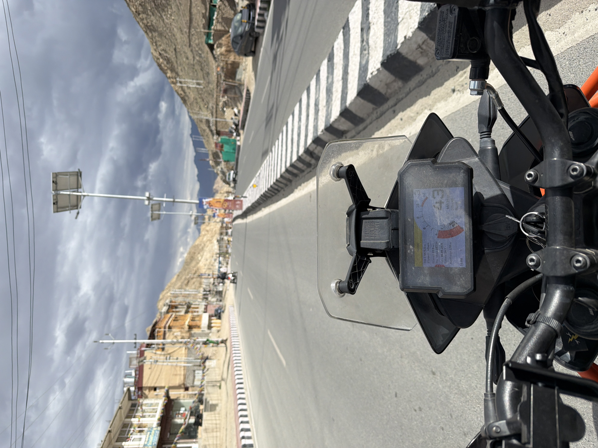 The image is a first-person perspective from a motorcycle in Leh, India, on a cloudy day. The motorcycle's dashboard displays the speed as 43, trip distance as 4475.5 km, and the date as 06.09.2026. The road is paved and appears to be a main thoroughfare, with buildings lining the left side and a mountainous, arid landscape on the right. Several vehicles, including a car and a scooter, are visible on the road. Solar-powered streetlights are present along the roadside. The sky is overcast with dark clouds.