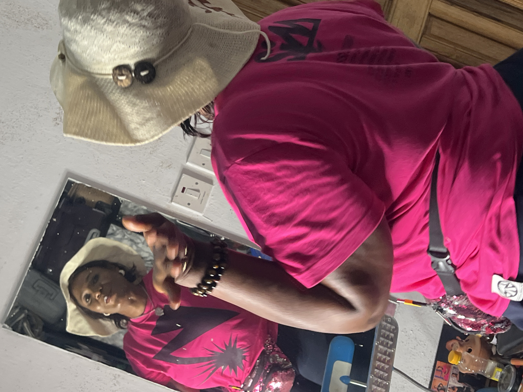 A woman wearing a bright pink t-shirt and a wide-brimmed straw hat is standing in front of a mirror, taking a selfie. The hat is decorated with two brown wooden beads. She is also wearing a black belt with a fanny pack that has a sequined design. In the mirror, her reflection shows her face, which is illuminated, and her extended left hand with jewelry on her wrist. She is wearing earrings and a necklace. The background is a plain wall, with a light switch visible on the right. The scene appears to be indoors. There is a water bottle and a small brown object on a shelf to the left. The image does not contain any information that would allow it to be located in Jos, Nigeria.