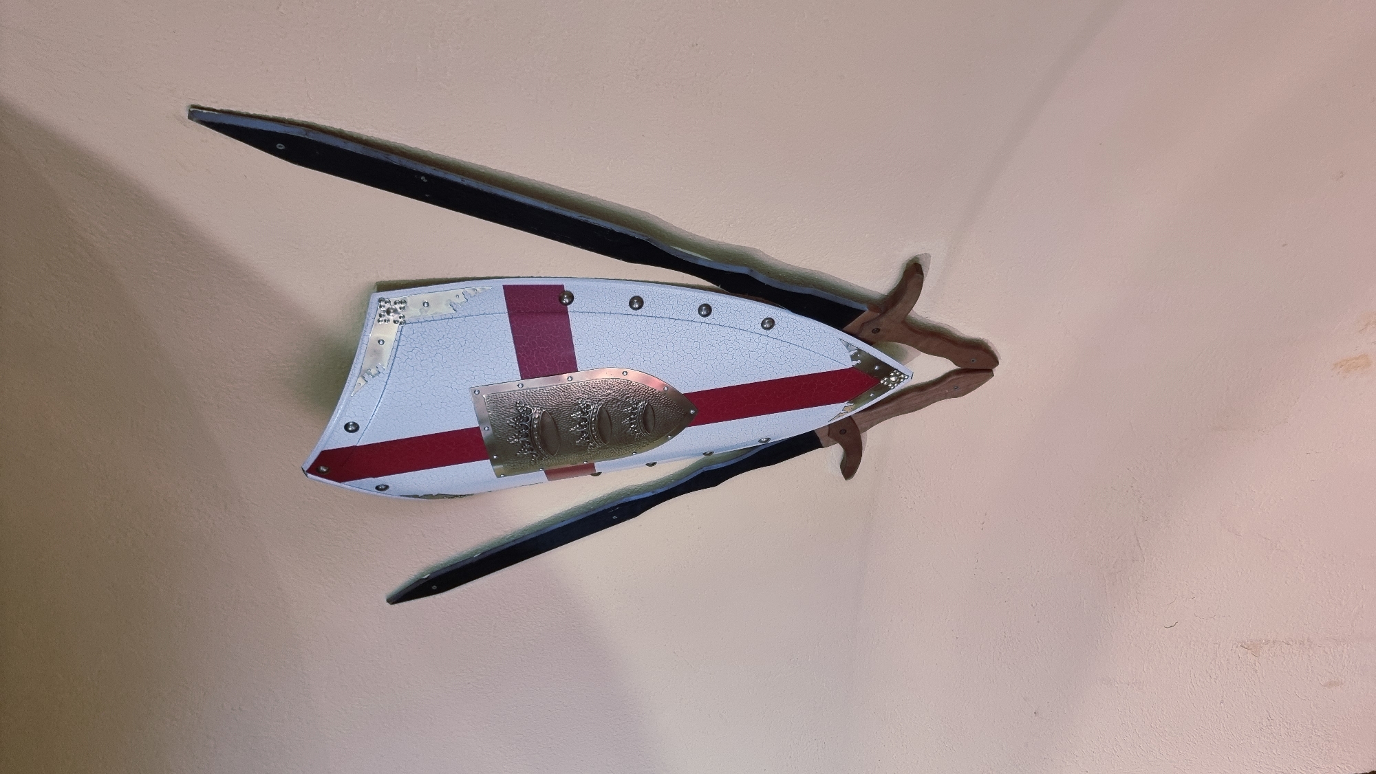 A decorative display of two swords crossed behind a shield is mounted on a textured, pale peach-colored wall. The shield is white with a red cross and is adorned with a smaller, ornate metal crest featuring three crowns. The swords have dark blades and wooden handles. The entire arrangement casts a shadow on the wall. The scene appears to be indoors, possibly in a historical building or museum in Besalú, Spain, given the style of the weaponry and shield.