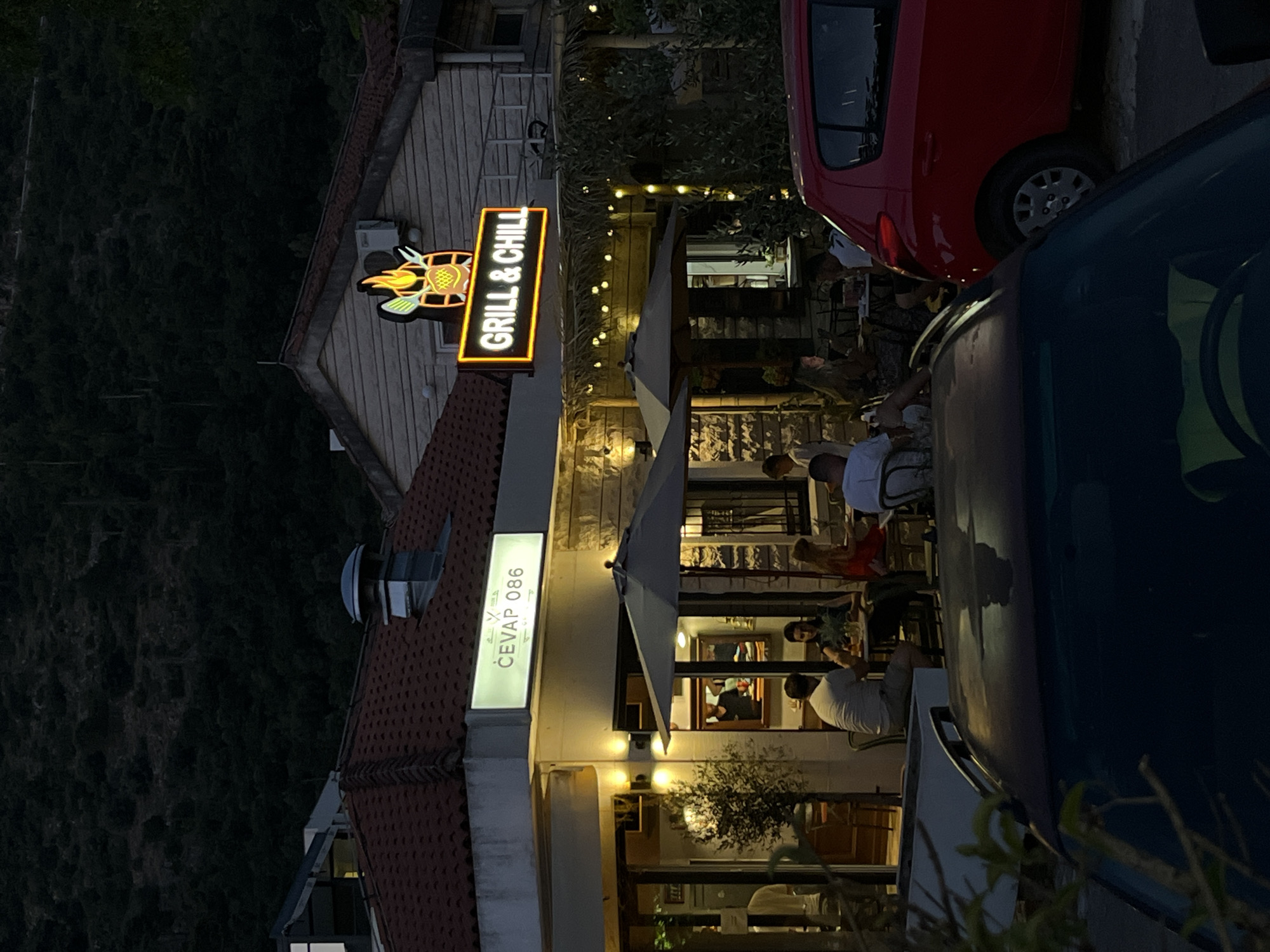 The image shows a street scene in Budva, Montenegro, at dusk. A restaurant with two signs, one reading "ČEVAP 086" and the other "GRILL & CHILL," is visible. Several people are seated at tables outside the restaurant, under umbrellas. A red car is parked to the right of the restaurant. The background features a dark, tree-covered hillside.