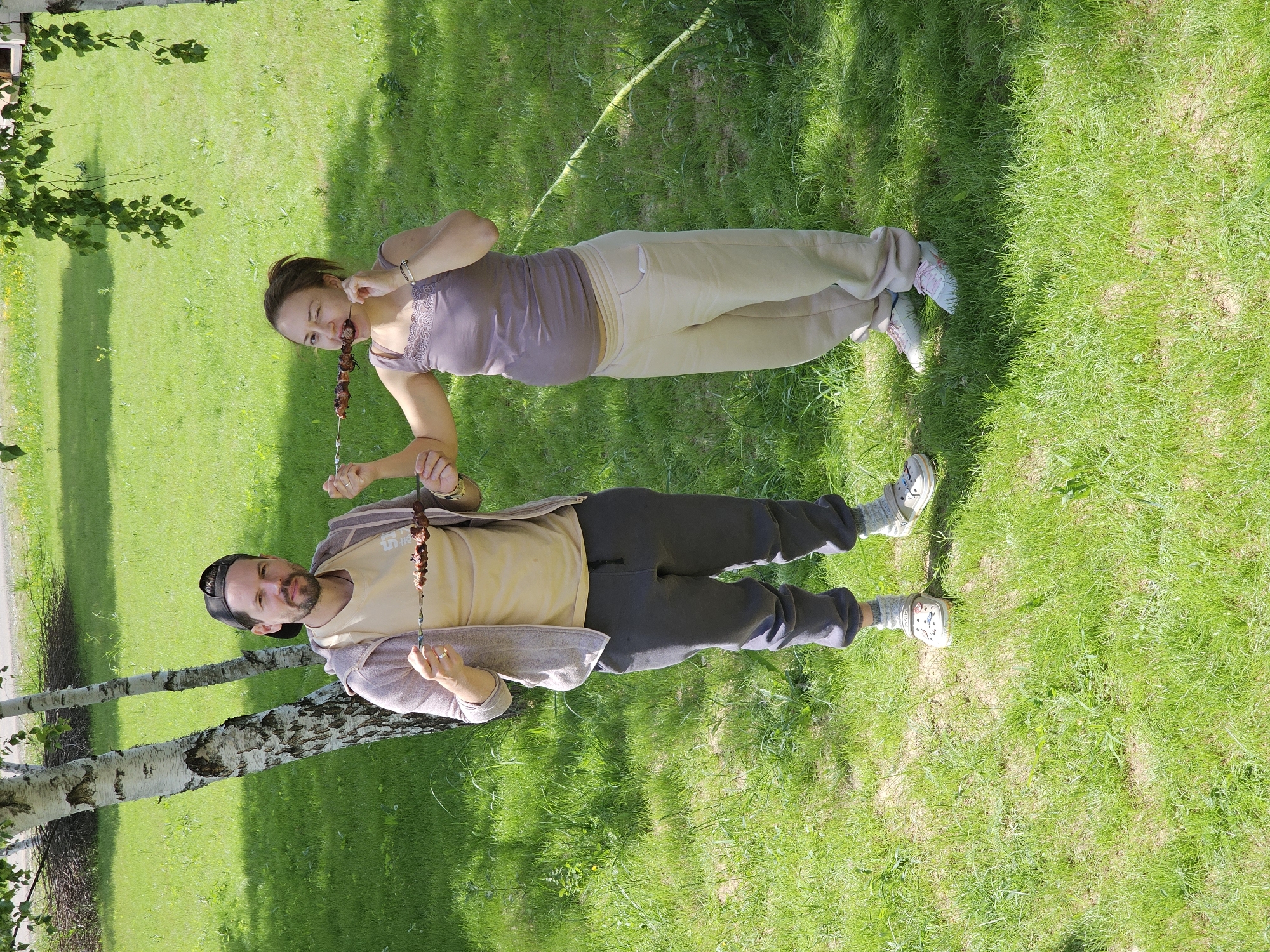 The image captures a man and a woman standing on a lush green lawn, both holding skewers of grilled meat. The woman, positioned higher in the frame, wears a brown tank top and beige sweatpants, and she is in the act of eating from the skewer. The man, lower in the frame, sports a tan shirt, gray sweatpants, and a black baseball cap. He is also holding a skewer. The background features birch trees and well-maintained grass, suggesting a park or recreational area. The location is specified as Мышкино, Russia.