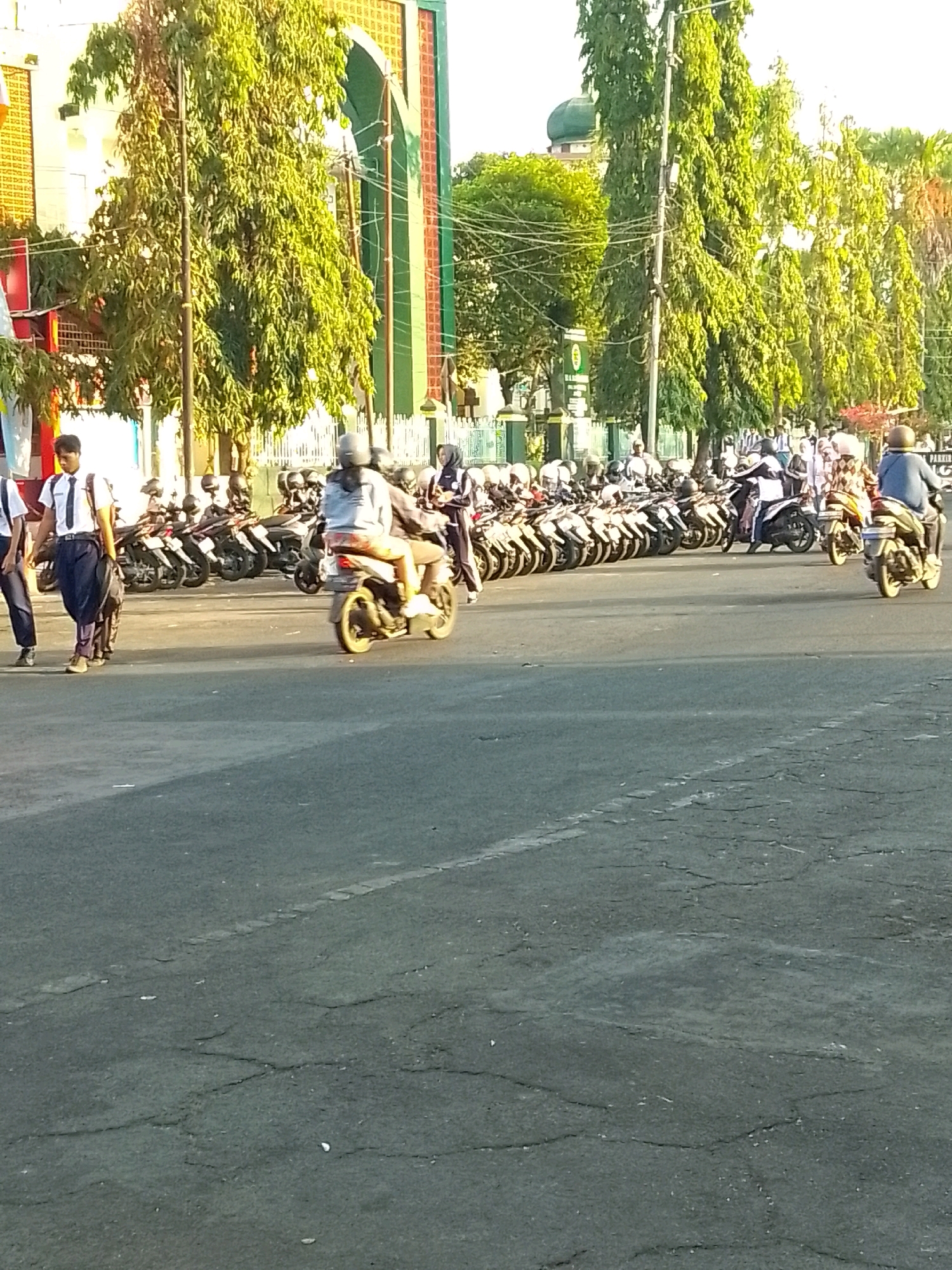 The image captures a street scene in Jember, Indonesia, with a prominent line of parked motorcycles on the left. Several individuals, some appearing to be students in uniform, are walking or standing near the motorcycles. In the background, a building with green and orange architectural features and a dome is visible, possibly a religious structure. Tall trees line the street, casting shadows and contributing to the bright, sunny atmosphere. A few people are riding motorcycles, with one on the far right appearing to be a man wearing a helmet and blue shirt. The road surface shows signs of wear and tear, with visible cracks. The overall impression is of a busy street with a mix of pedestrians and vehicles, set against a backdrop of urban architecture and vegetation.