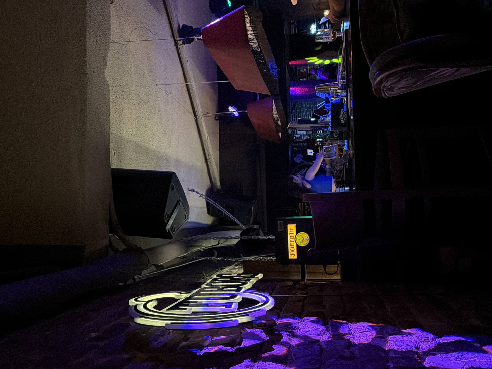 The image shows the interior of a bar, likely in Sofia, Bulgaria, based on the Cyrillic lettering on the illuminated sign on the floor. The foreground is dominated by the glowing white and blue neon sign, which is resting on a brick or cobblestone surface, with the blue light creating purple reflections on the stones. A dark metal pipe runs along the left edge of the frame, and a large black speaker is visible next to the wall. To the right of the sign, a Jägermeister promotional display sits near the bar area.
In the background, a bartender in a blue shirt is serving drinks, with shelves of liquor bottles and illuminated signage above them. Red awning-like structures hang from the ceiling, and other light sources create a dimly lit environment. There are a few customers visible at the bar, but most details are obscured by shadows.