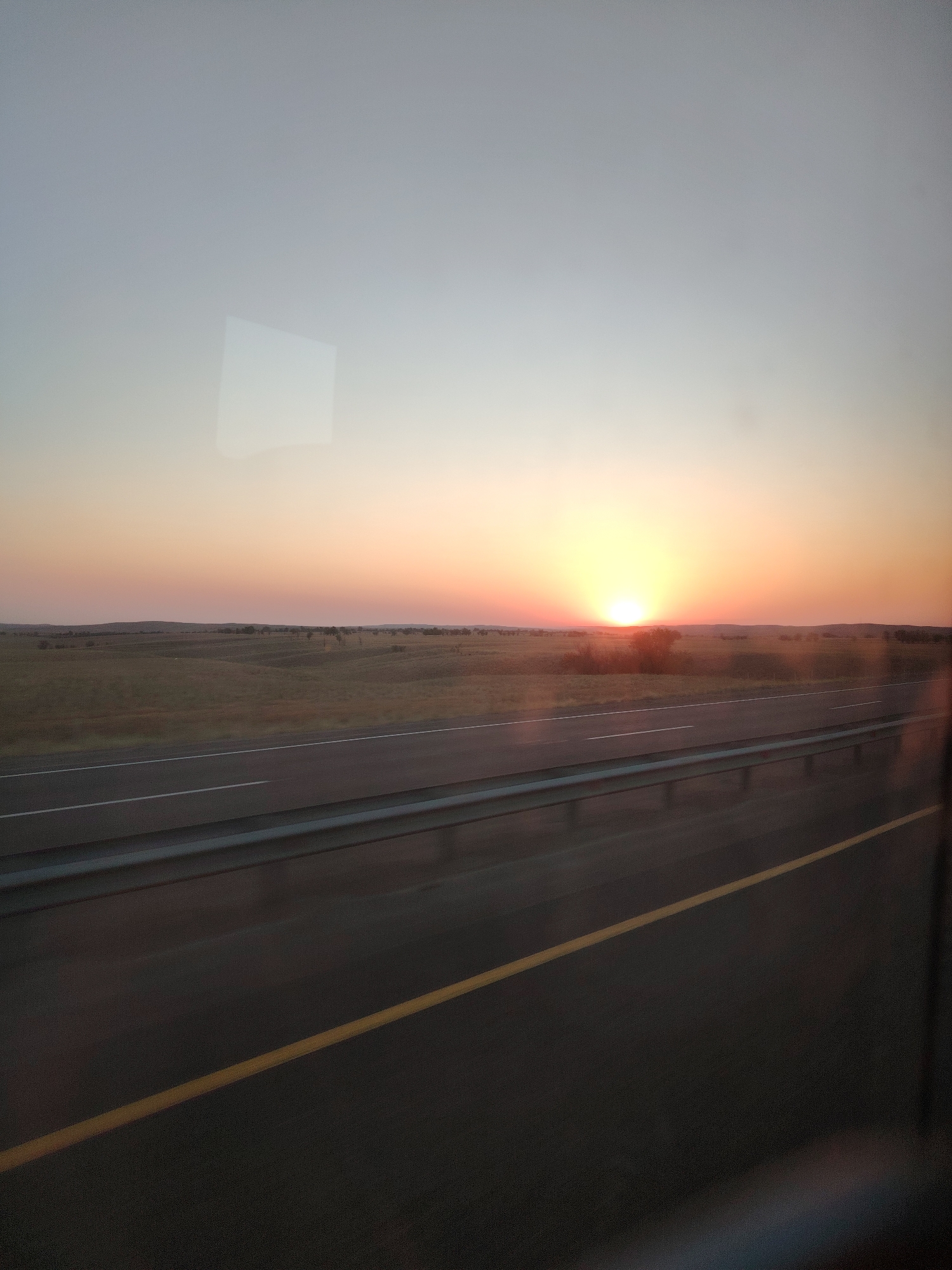 This image captures a sunset over a vast, grassy plain with a highway in the foreground, viewed from what appears to be inside a vehicle. The sky transitions from a pale blue at the top to vibrant hues of orange and yellow near the horizon, where the sun is setting behind a low range of hills. The landscape is characterized by dry, golden-brown grass dotted with sparse vegetation. A two-lane highway runs horizontally across the middle of the frame, separated by a guardrail. The road surface is dark asphalt with white and yellow lane markings. A subtle rectangular reflection, possibly from a windowpane, is visible in the upper left quadrant of the sky. The overall impression is one of open space and natural beauty, suggesting a rural or semi-rural setting. The location is identified as Samsy, Kazakhstan.