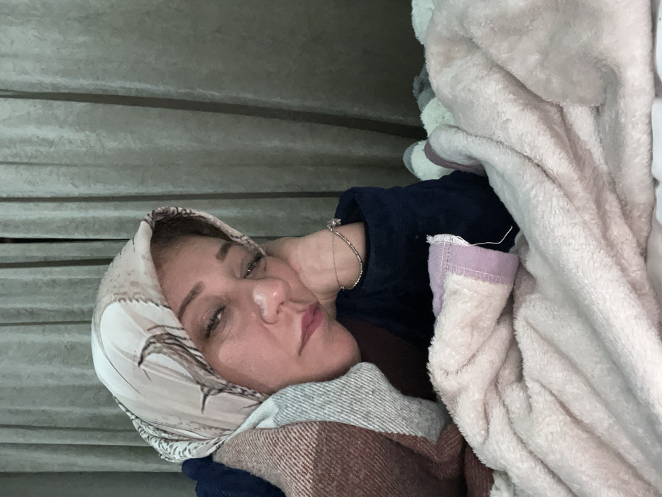 A woman with light skin is resting, her head covered in a patterned scarf. She's wrapped in a maroon and grey blanket, a blue garment peeking out from beneath. A silver bracelet adorns her wrist. Fluffy, light-pink material engulfs her right side. A textured, metallic-grey wall is visible in the background. The woman appears relaxed, with closed eyes and a serene expression.