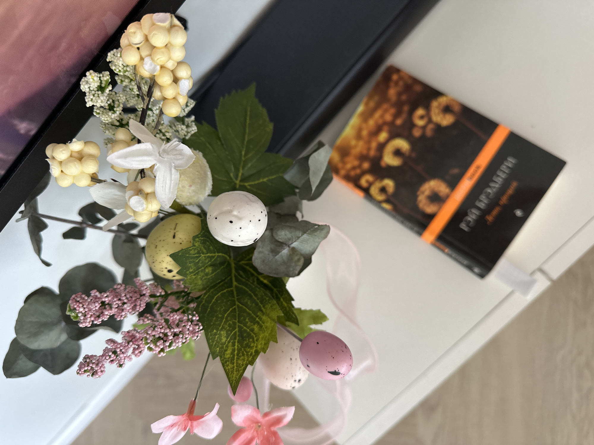The image shows a close-up, high-angle view of a decorative Easter arrangement and a book placed on a white surface, likely a table or shelf. The arrangement consists of artificial flowers, leaves, and speckled Easter eggs in pastel colors (white, yellow, and pink). A light pink ribbon is intertwined within the arrangement.
Next to the arrangement is a book with a black cover and an image of what appear to be dandelion seed heads illuminated with a warm, orange glow. The book title is partially visible in Cyrillic script.
The background is out of focus, but it appears to include part of a television screen or monitor with a frame, and hints of flooring below the white surface. The setting appears to be indoors.
There is no evidence to suggest that this image was taken in Kharnavy, Russia.