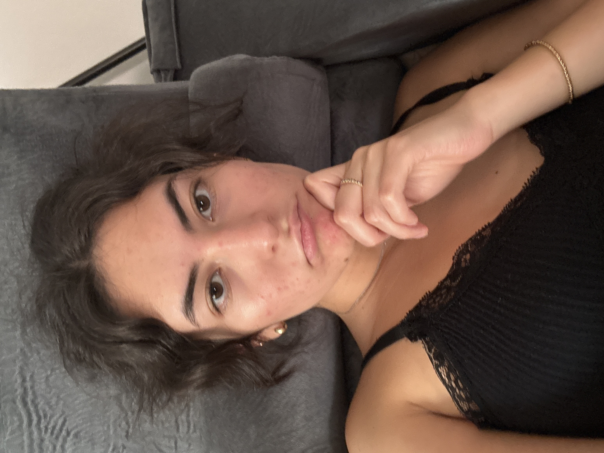A woman is lying on a grey couch. She has brown hair and is wearing a black top with lace detailing. She is wearing a gold ring on her finger and a gold bracelet on her wrist. The location is unknown and cannot be determined from the image.