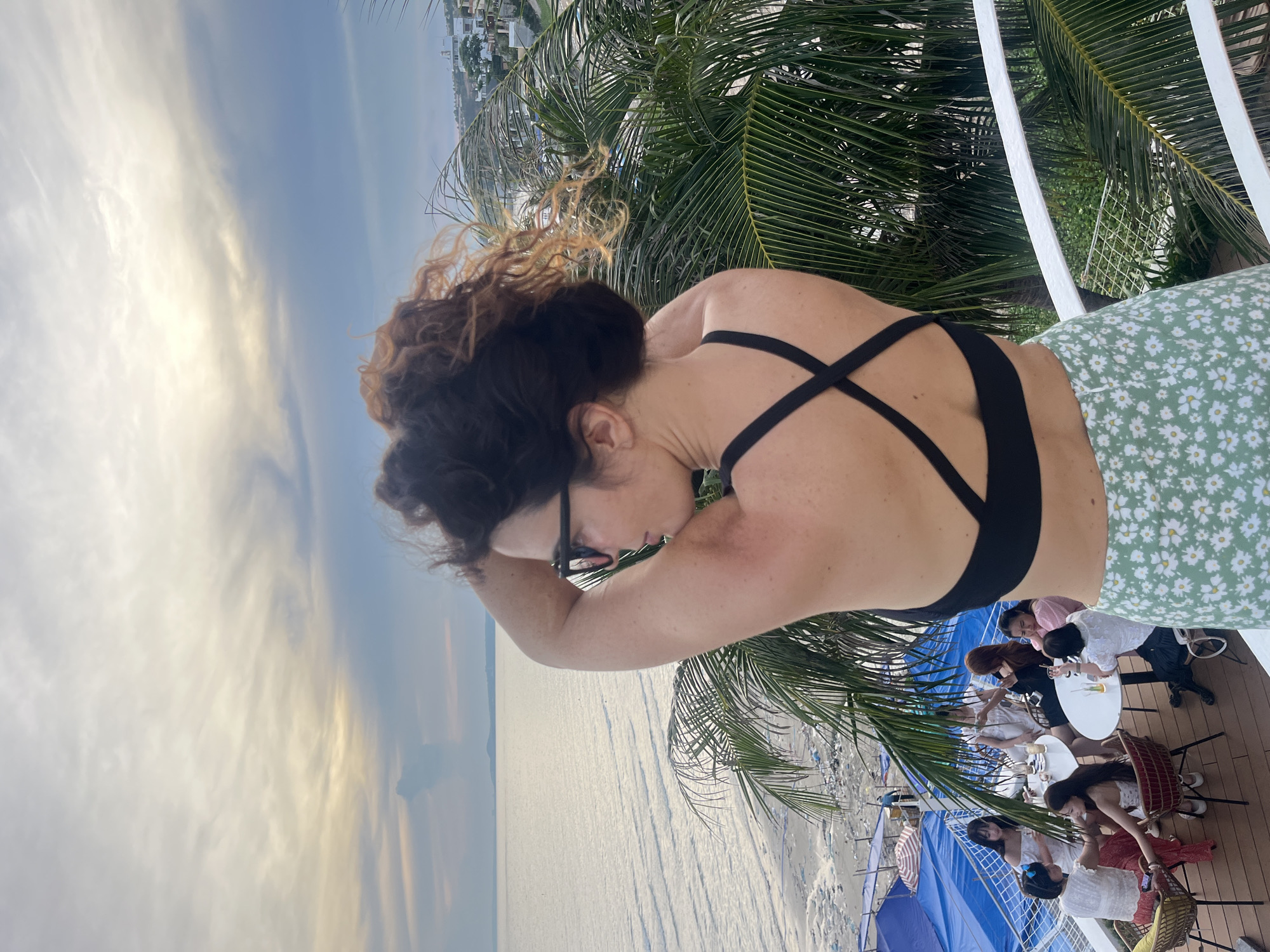A woman with dark curly hair and sunglasses is standing on a balcony in Mũi Né, Vietnam, facing away from the camera and looking out at the ocean. She is wearing a black sports bra and a green and white floral skirt. Her arms are raised, and she appears to be adjusting her hair. Below the balcony, a group of people are seated at tables, seemingly enjoying drinks. Palm trees are visible in the foreground and background, and the ocean stretches out to the horizon under a cloudy sky. The location appears to be a restaurant or bar with a view of the beach.
