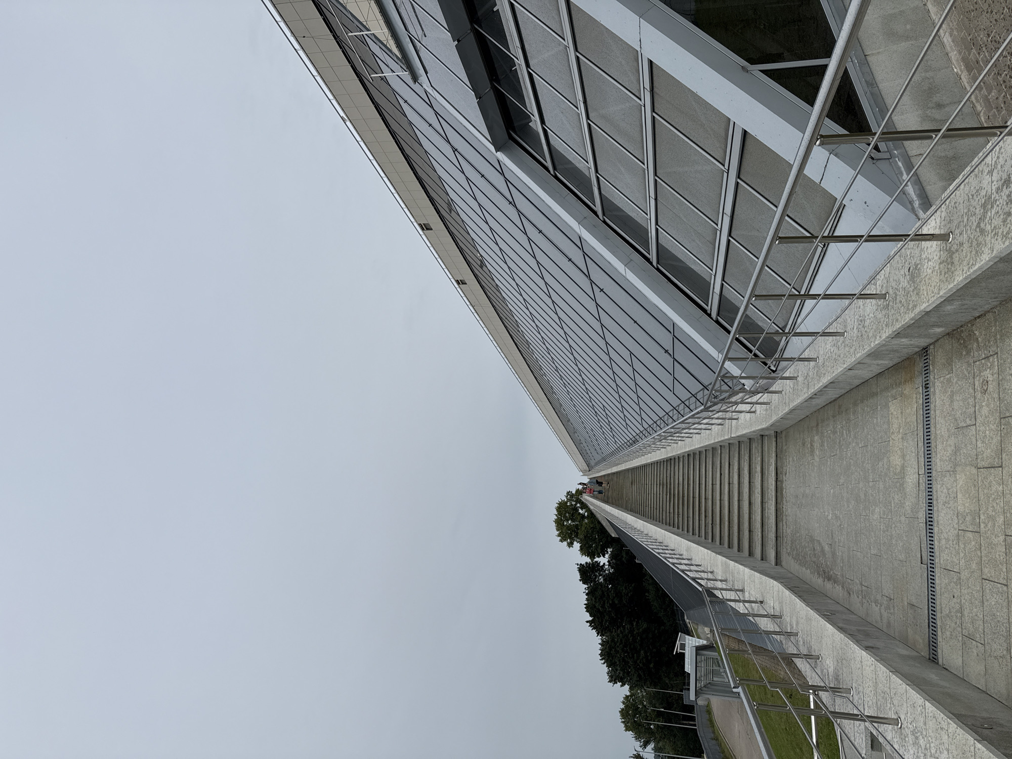 The image depicts a long, wide staircase leading upwards, flanked by a modern building with a slanted glass facade. The sky is overcast and gray. Three people are visible walking up the stairs in the distance. The staircase and building are constructed from concrete and metal, with a paved walkway at the base of the stairs. The environment appears to be an outdoor public space. Based on its architectural style and the presence of a large, modern structure with extensive glass, this could be a part of the Kaluga Drama Theater in Kaluga, Russia, which features similar contemporary design elements and an imposing staircase.