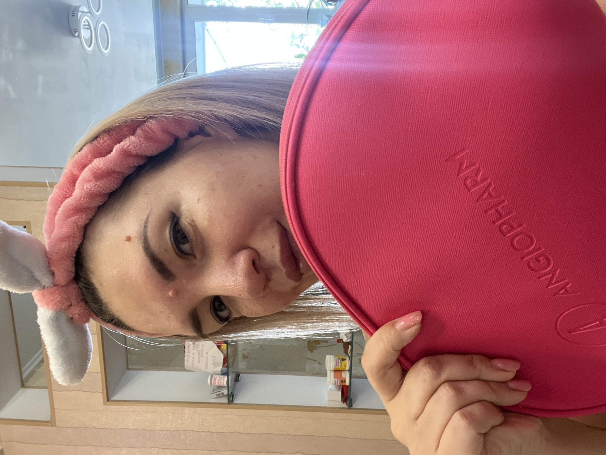 A woman is holding a pink bag in front of her face. She is wearing a pink bunny ear headband. The background shows shelves with bottles and a window. The image is a selfie taken indoors. There is no information in the image to determine the location in the city of Sochi, Russia.