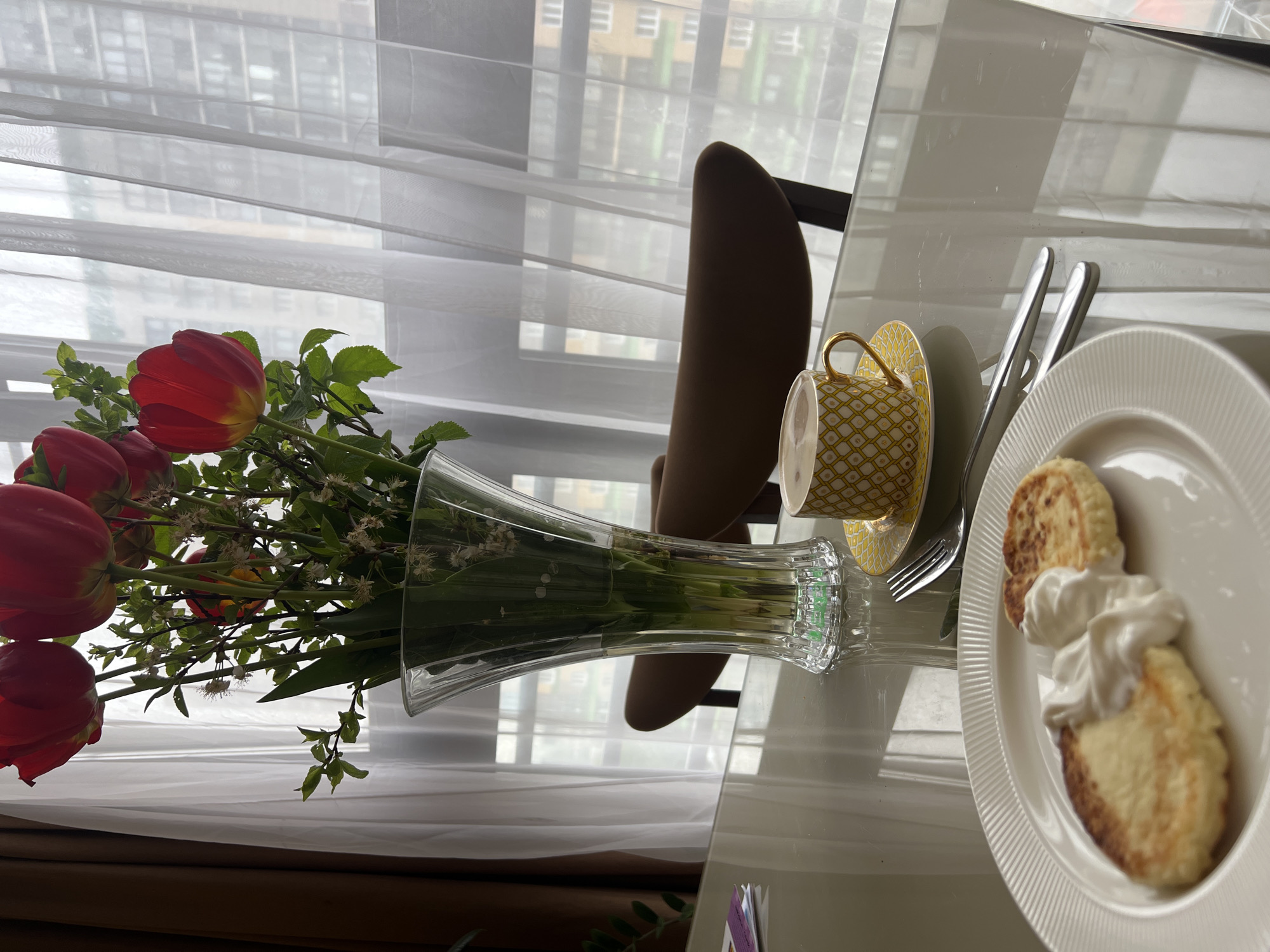 A table setting with a vase of red tulips and a plate of syrniki with sour cream sits beside a window, likely in Khimki, Russia. A yellow patterned teacup and saucer are next to the plate. Silverware is partially visible between the cup and plate. The setting is likely indoors. A chair is partially visible behind the table setting. The background shows a building through the window.
