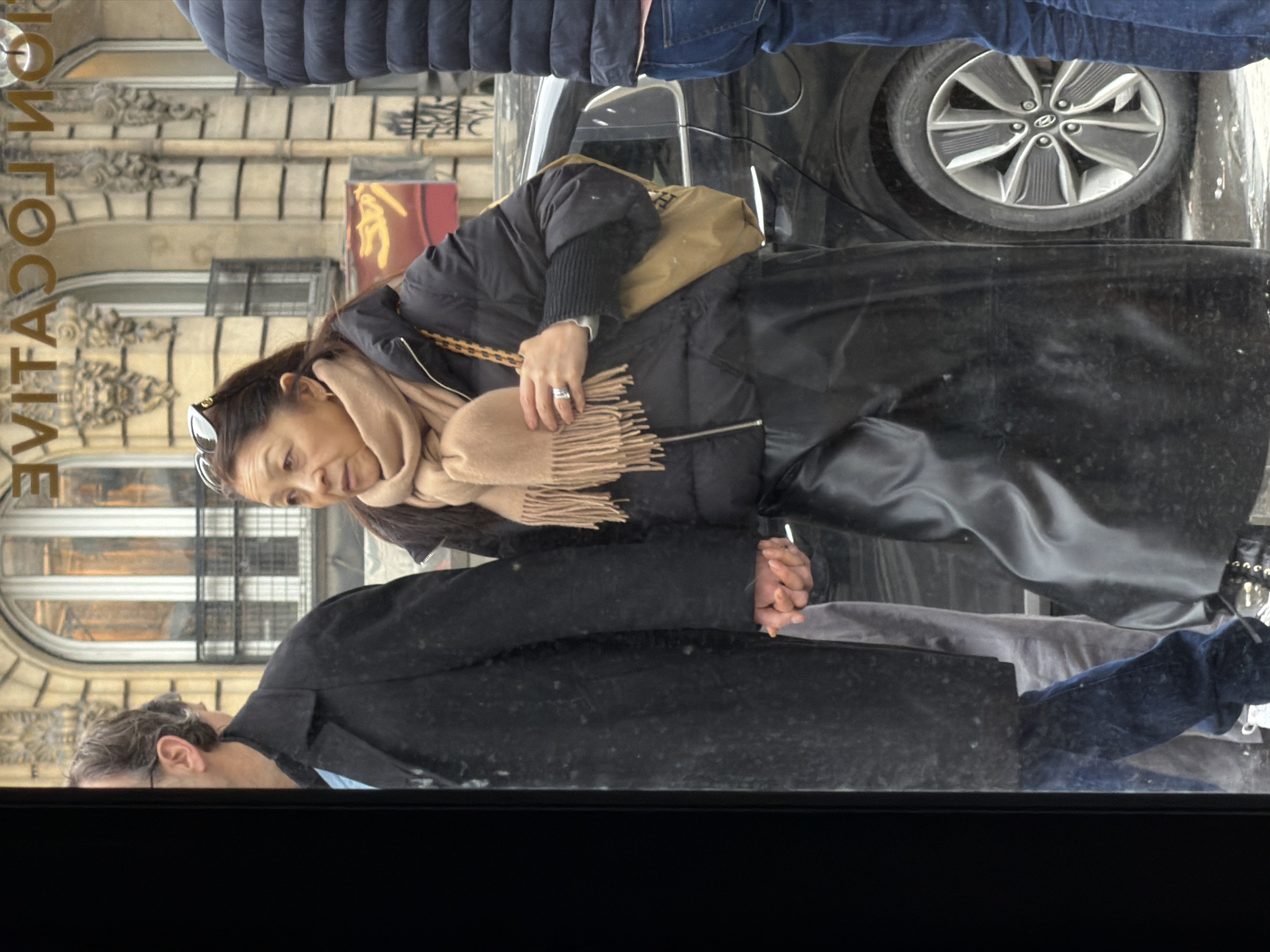 The image depicts a reflection in a window, showing people and elements of the Parisian urban environment. A woman wearing a black puffer jacket, a long black leather skirt, and a beige scarf is captured holding hands with a man wearing a dark coat. The reflection shows elements of the building across the street, including arched windows and architectural details with the vertically oriented lettering of "LOCATIVE" visible, suggesting a business name. Above the woman is a reflection of the lower portion of a person wearing jeans and a dark blue puffy coat, along with a car with an alloy wheel. The overall impression is of a busy Parisian street scene captured indirectly through a window reflection.