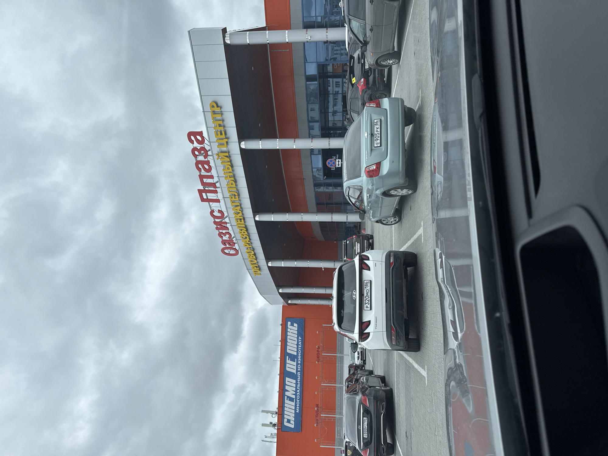The image shows a view of the "Oasis Plaza" shopping and entertainment center in Beloyarsky, Russia, captured from inside a vehicle. Several parked cars are visible in the parking lot in front of the building. The license plates on the vehicles include E888PT 186, P320MO 186, and E905 PK 86. A sign for "Cinema de Luxe," a multi-screen 3D cinema, is visible on the building's facade. The sky is overcast.