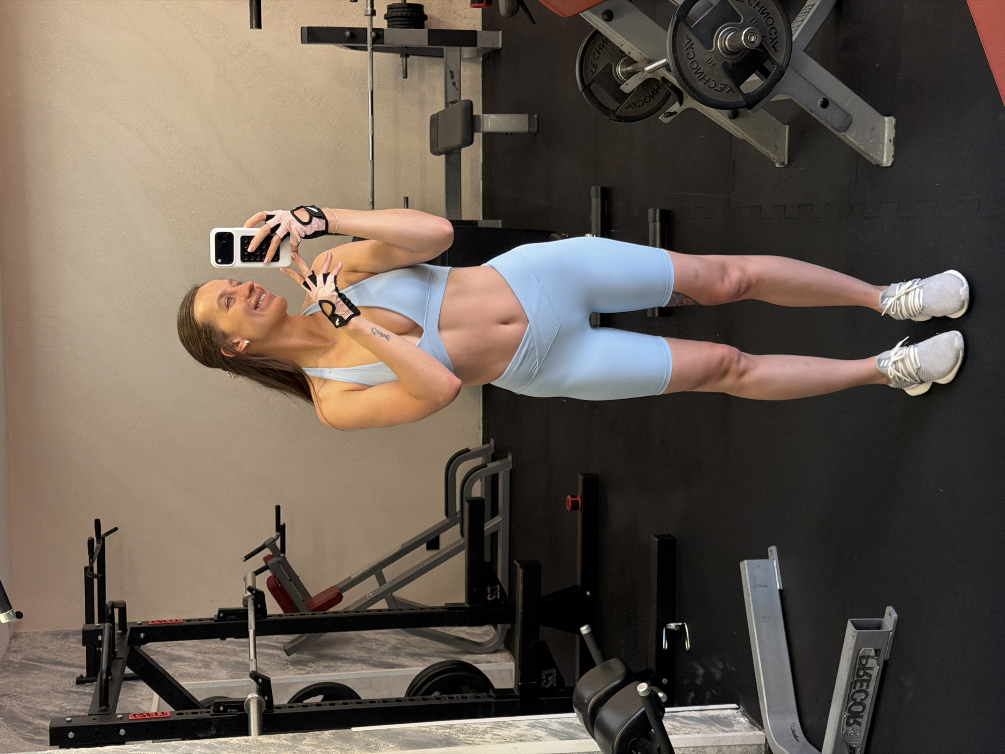 A woman in a light blue athletic set, consisting of a sports bra and high-waisted shorts, stands in a gym and takes a selfie. She is wearing pink and black weightlifting gloves on both hands and grey athletic shoes. She has long brown hair and is smiling. The gym appears to be equipped with various weightlifting machines and racks, including a pull-up bar and benches. The floor is black, and the walls are a light beige color. The location is identified as the city of Budva, Montenegro.