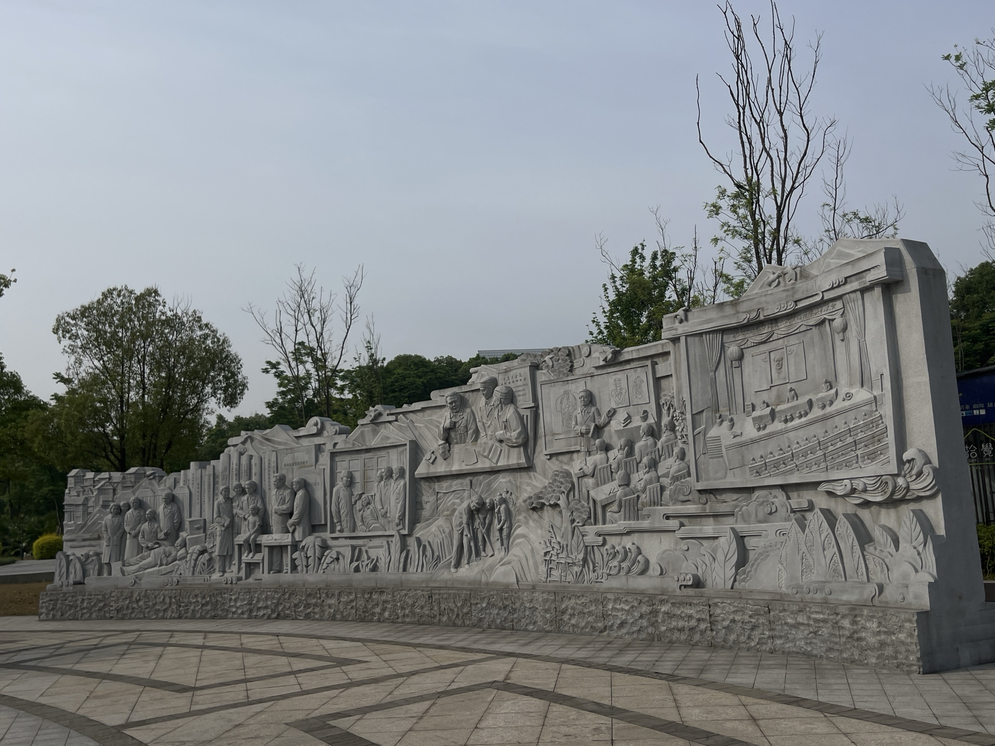 The image depicts a large stone relief sculpture located in the Chongqing High-tech Zone, China. The sculpture is a long, curved wall displaying various scenes in intricate detail, seemingly depicting historical or cultural events. The relief features numerous figures engaged in different activities. There are trees in the background, some bare and some with green foliage. The ground is covered in a light-colored tiled pavement with a geometric pattern. The sky is overcast and a light gray color.