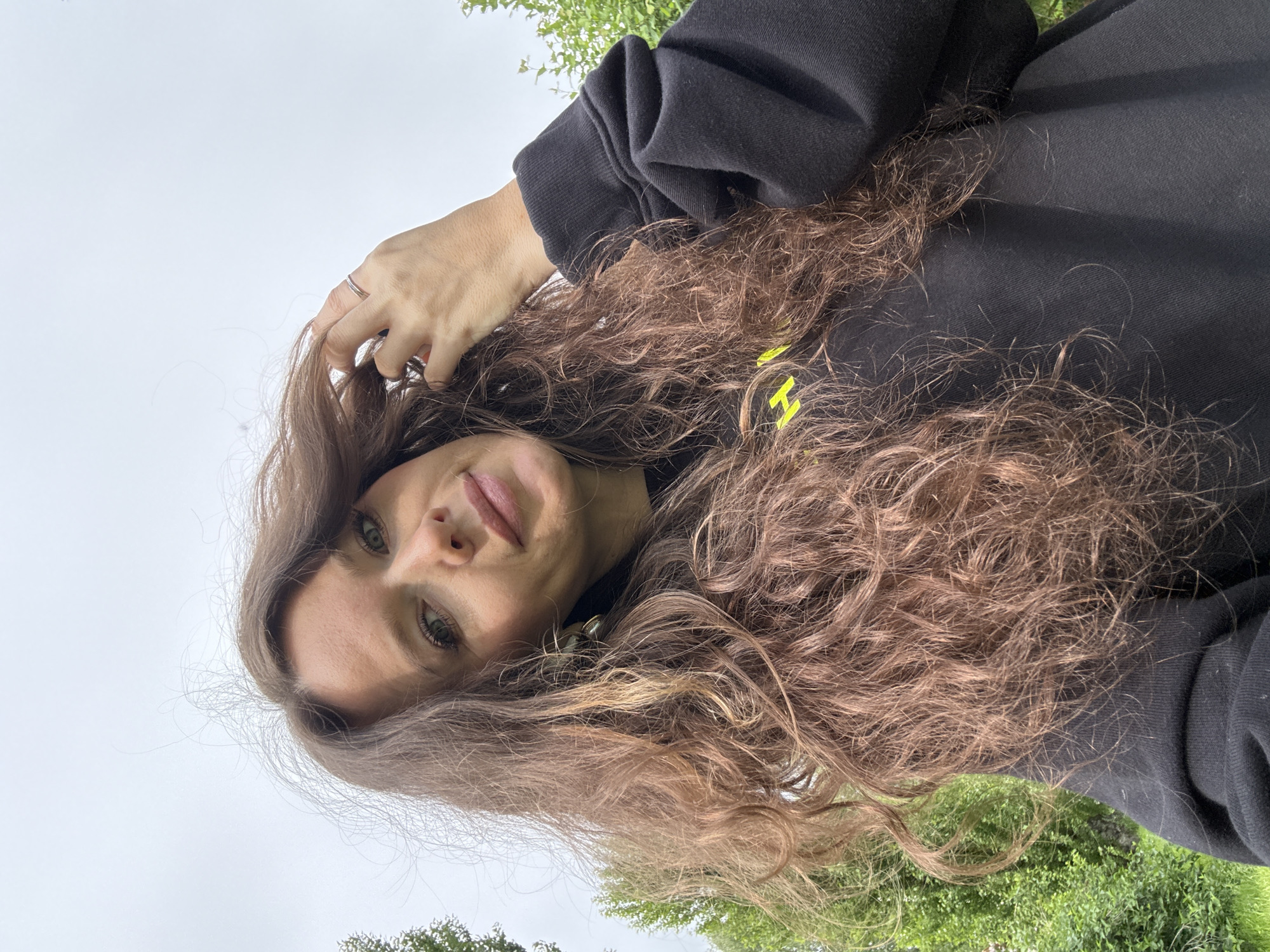 A woman with wavy brown hair and green eyes is looking into the camera. She has fair skin and light pink lipstick. She's wearing a black sweatshirt. Her left hand is raised and touching her hair. The background shows grass and trees, and a cloudy sky. The location appears to be outdoors, potentially a park. Moscow, Russia is not evident in this image.