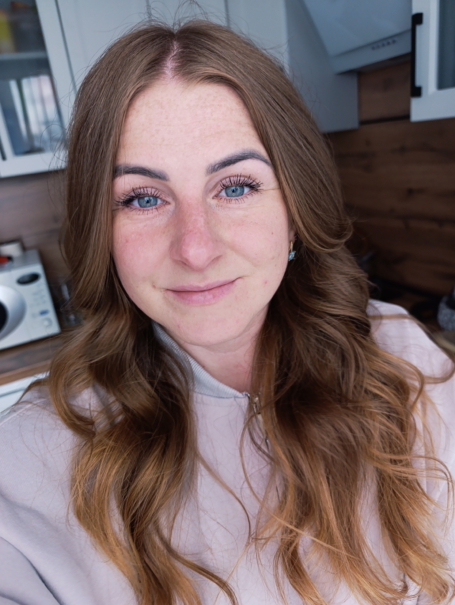 A close-up, eye-level shot depicts a woman with light-brown, wavy hair and fair skin covered with freckles. She has blue eyes enhanced with mascara and a slight smile. The background consists of a wooden counter, white cabinets, and a white appliance on the counter. The setting suggests an interior scene. There is no evidence of location in the city of Unknown, Unknown.