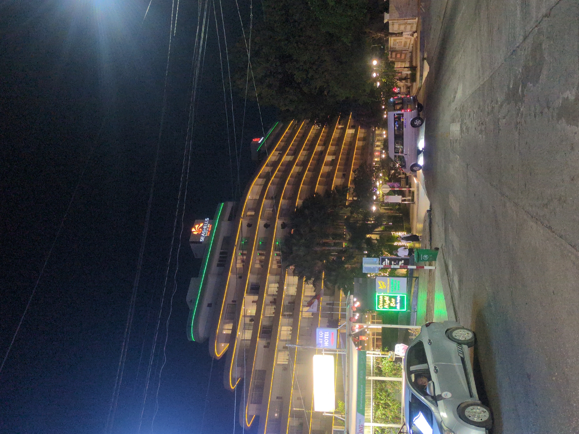 The image shows an exterior view at night, featuring the Seashells Hotel & Spa Phú Quốc. The hotel is lit with warm-toned lights along its curved balconies and a green light accentuating its height. In front of the hotel, various signs are visible, including one for "Ocean Night Bar." A silver car is parked on the paved area adjacent to the signs. Wires are strung above, adding to the urban setting. People are visible in the background near the hotel entrance. A white van is also parked nearby. The location is Phú Quốc, Vietnam.