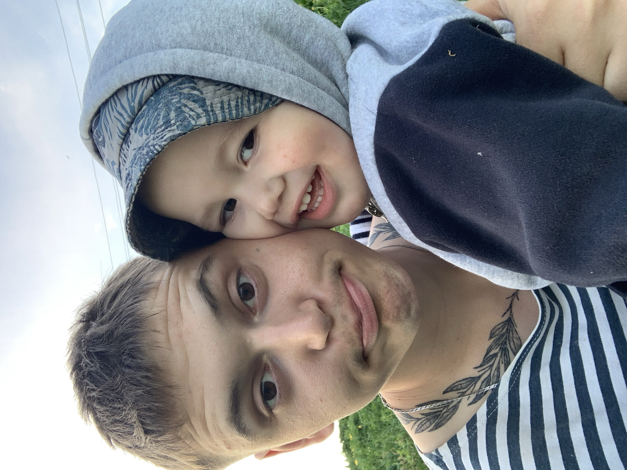 A selfie shows a man and a young child, possibly in the city of Фоки, Russia, due to the user's query. The man has short, light brown hair, brown eyes, and appears to be wearing a striped shirt with a leaf-like tattoo on his neck. The child, wearing a gray hoodie and a patterned cap, is nestled close to the man, smiling with visible teeth. They are outdoors, with a glimpse of the sky and greenery suggesting a park or natural area. The lighting indicates it is daytime.