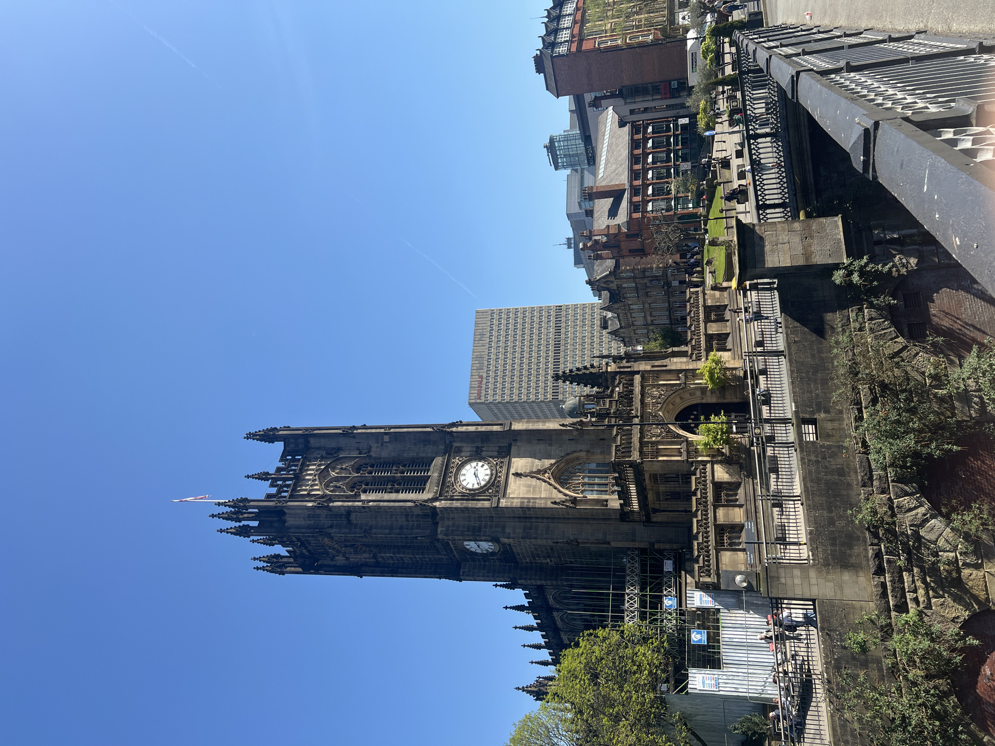The image shows Manchester Cathedral in Salford, United Kingdom. The cathedral's Gothic architecture is prominently displayed, with a tall tower featuring two visible clocks. Buildings of varying heights and architectural styles are visible in the background, indicating an urban setting. A black metal railing is in the top right corner, possibly part of a bridge or walkway. Some people are walking along a pathway adjacent to the cathedral. Construction or maintenance work seems to be underway with temporary barriers and scaffolding present. The sky is clear and blue.