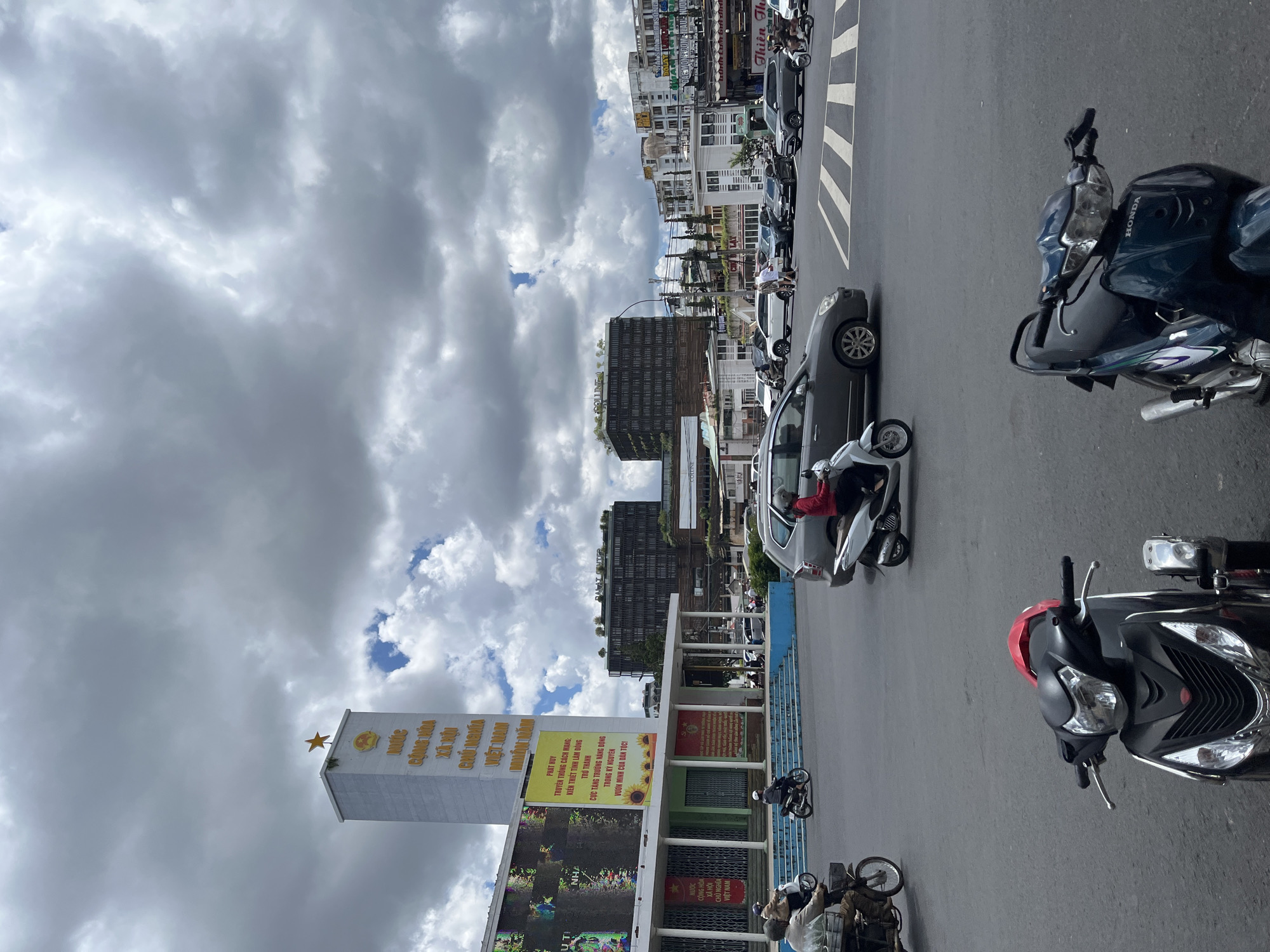 The image depicts a street scene in Da Lat, Vietnam, likely at a busy intersection. In the foreground, two parked motorcycles are visible, one black with red accents and the other dark blue. The midground shows a grey sedan in motion, with a rider on a scooter behind it. Further back, more vehicles, including cars and scooters, are navigating the intersection. On the left side of the image, a tall, white tower with a golden star at its apex stands prominently. This tower features Vietnamese text in yellow. Adjacent to the tower is a building with a large, illuminated screen. Beyond these structures, several multi-story buildings, some with modern architecture and greenery on their rooftops, form the urban landscape. The sky is filled with dramatic, grey clouds, suggesting overcast weather. The overall impression is a bustling city intersection in Da Lat.