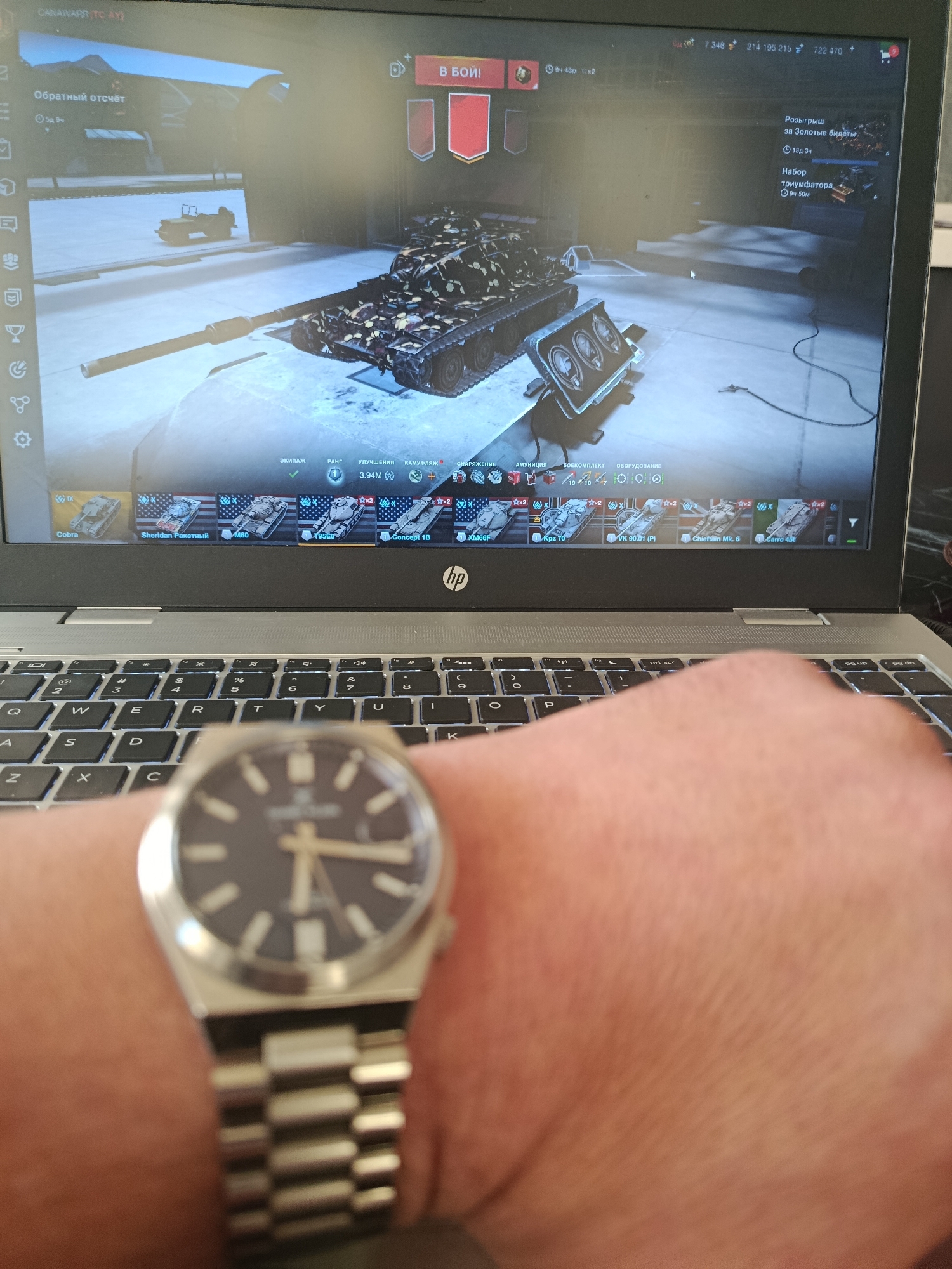 The image displays a laptop screen with a video game, likely World of Tanks, open. The game interface shows a tank in a garage or hangar setting. Various icons and text related to game statistics and features are visible. Below the laptop, a keyboard is partially visible. In the foreground, a person's wrist is adorned with a silver wristwatch, which is the primary subject of the image. The background is blurred, suggesting a focus on the watch and the laptop screen. There is no discernible information within the image to suggest the location is in Baku City, Azerbaijan.