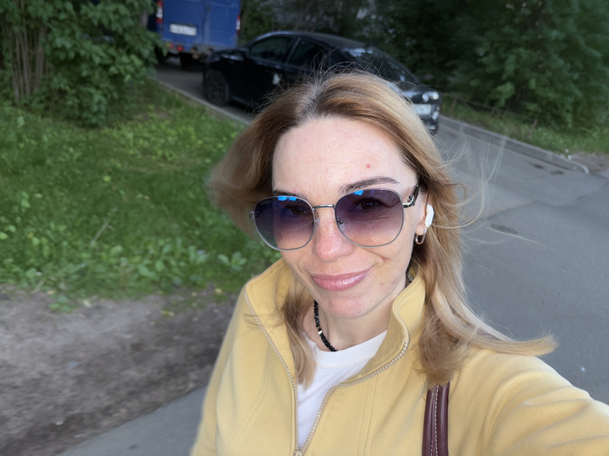 A woman with blonde hair and sunglasses is taking a selfie outdoors. She is wearing a yellow jacket over a white shirt and a black beaded necklace. A black car and a blue truck are visible in the background. The scene appears to be a street with trees and grass.