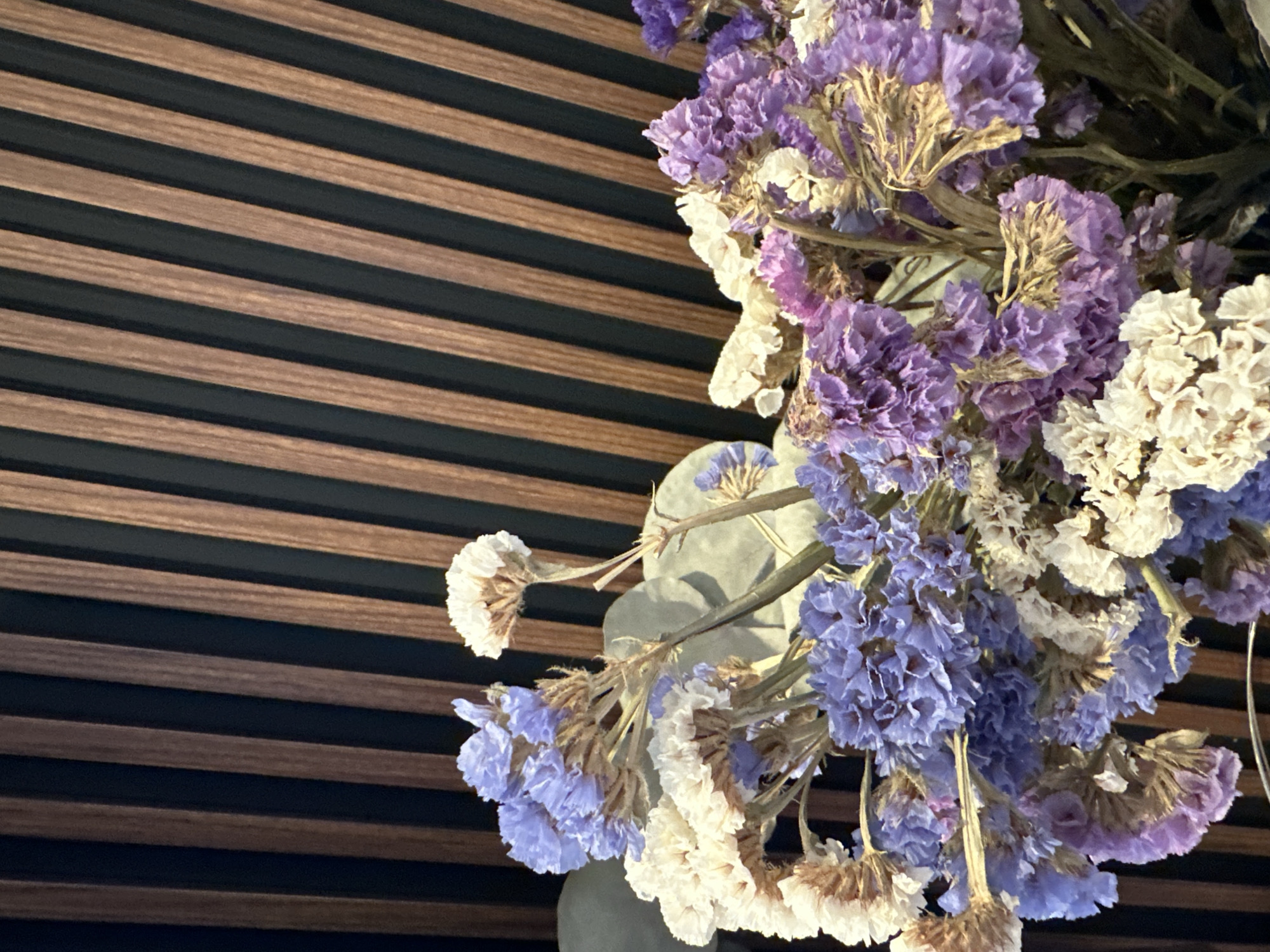 The image shows a close-up of a bouquet of dried flowers, primarily in shades of purple, blue, and white, set against a background of dark vertical wooden slats with thin black gaps. The flowers are small and clustered, with some stems and dried seed pods visible. The lighting is soft, highlighting the textures of the dried petals. There is no indication of any specific location within Minsk, Belarus.