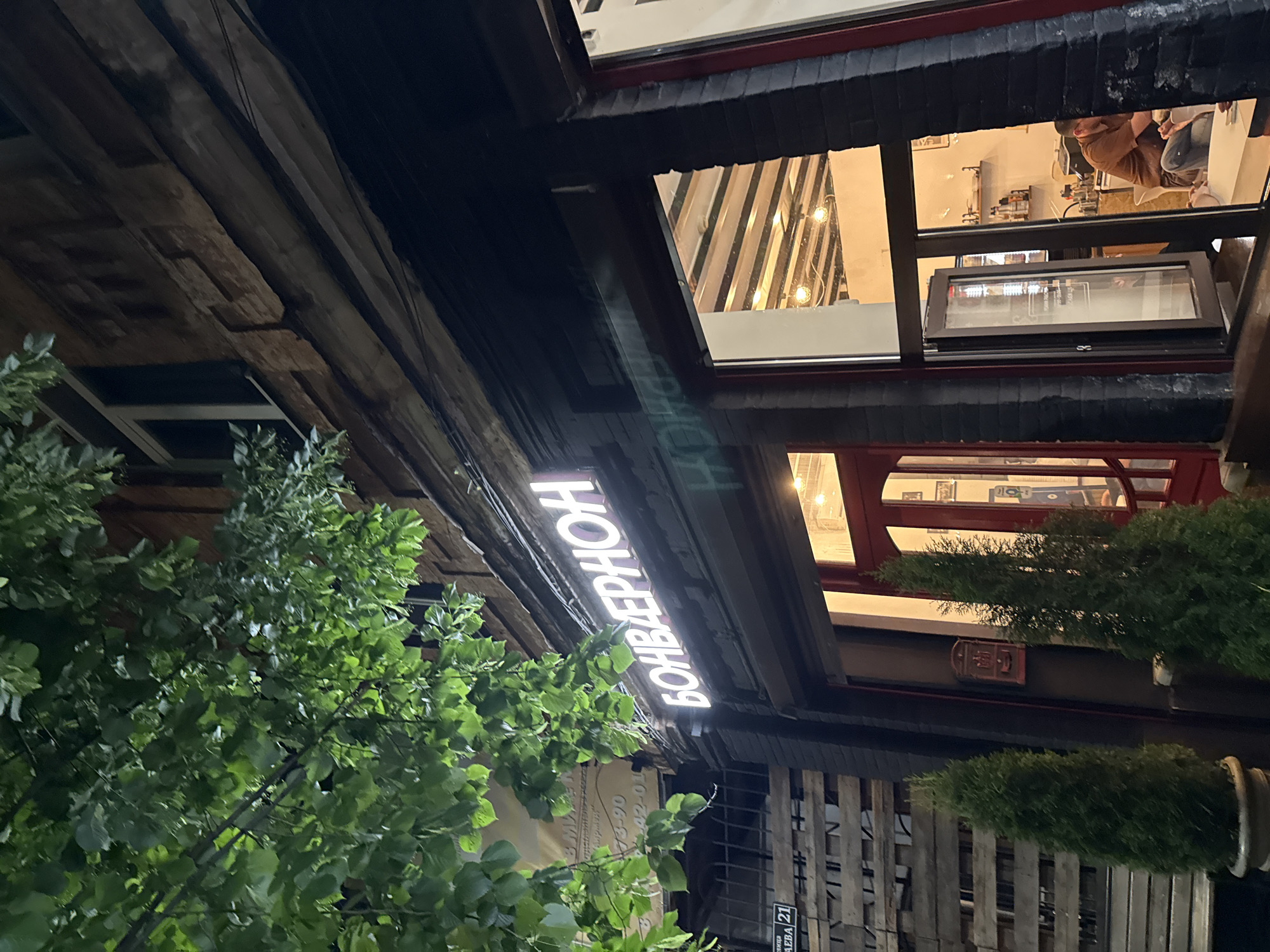 This media file captures an exterior view of a building at night, likely a commercial establishment. The building features a mix of rustic and modern architectural elements, with wooden beams and siding juxtaposed with large windows and a glowing neon sign.
The main subject of the image is the illuminated sign with Cyrillic text that reads "БОНВАЛЕРРНОН". The sign is white and positioned vertically against the dark wooden facade. In the background, through several large windows, interior lights are visible, suggesting that the establishment is open and operational. One window offers a glimpse of people inside, possibly staff or patrons, engaged in activities. The lighting in the interior appears warm and inviting.
Green foliage from a tree or bush is prominent in the foreground, partially obscuring the lower part of the building and the sign, adding a natural element to the urban scene. The overall atmosphere is nocturnal, illuminated by the artificial lights of the building and the sign, with no clear indication of the weather.
A small sign with the number "21" is visible at the bottom right, possibly indicating a street number or an address. The text on the neon sign is the most prominent text in the image. Given the location context of Vladikavkaz, Russia, the Cyrillic text is consistent with this. The time of day is clearly night.