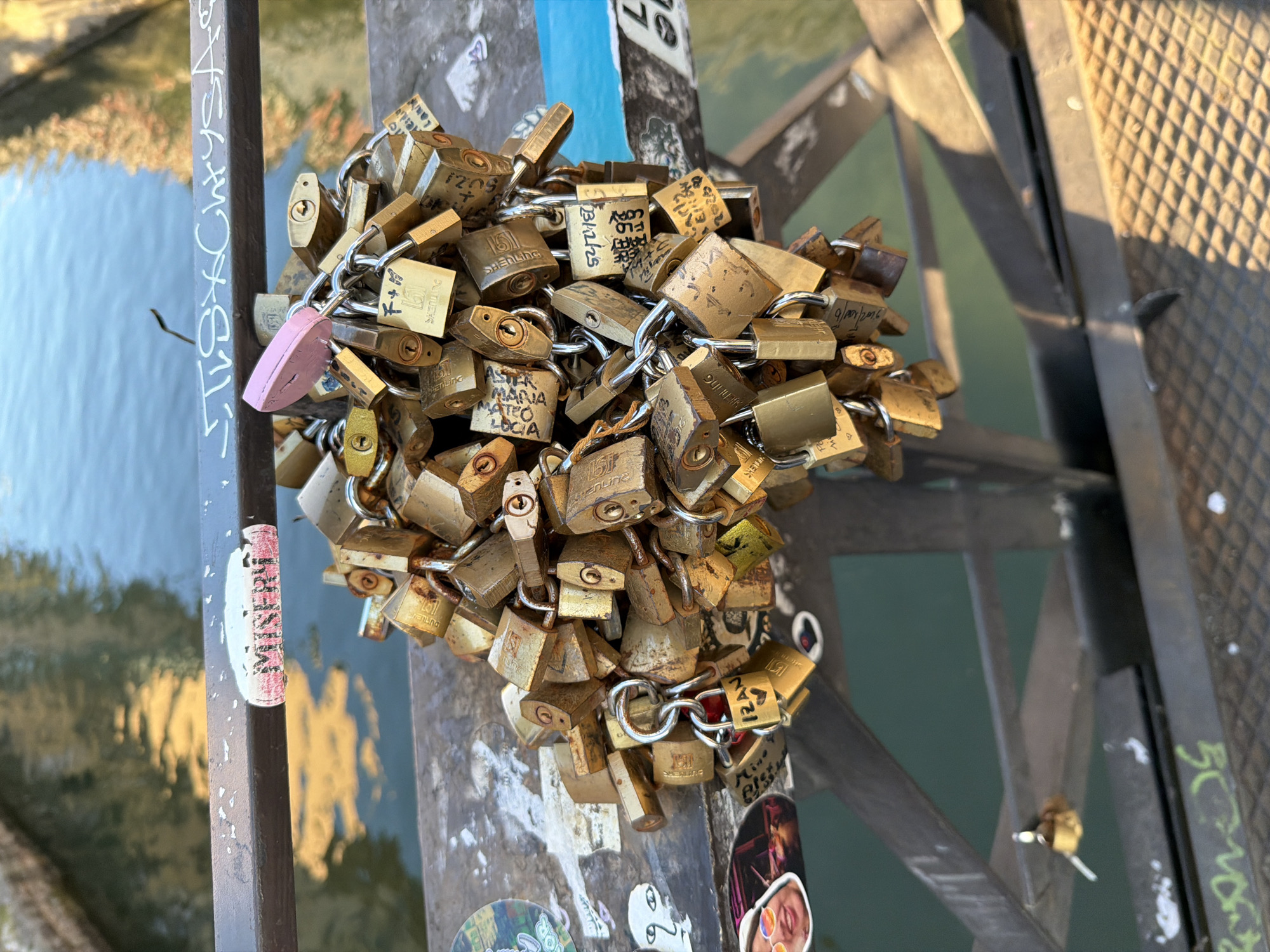 The image depicts a dense cluster of love locks attached to a metal support structure. The locks are primarily brass-colored, with one pink lock visible among them. Many locks have inscriptions, names, and dates. The support structure is covered in stickers and graffiti. Reflections of light and buildings can be seen in the water visible in the background. The location appears to be a bridge over water in Milan, Italy.