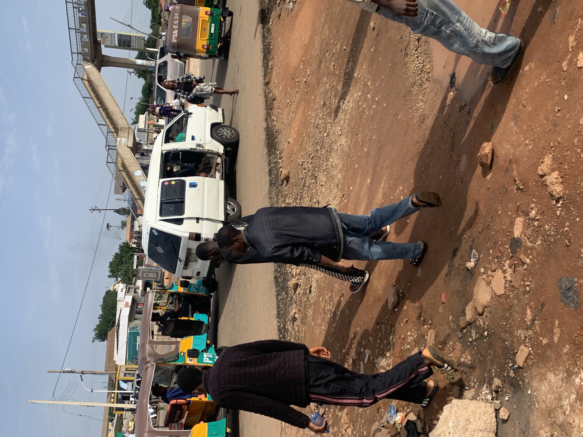The image depicts a street scene in Gura Top, Nigeria, featuring a mix of pedestrian and vehicular traffic. In the foreground, three men are walking on a dirt road. The man on the left, facing away from the camera, wears a dark quilted jacket and black track pants with pink stripes. He is holding a clear plastic bottle of water. The man in the middle, also walking away from the camera, is wearing a dark jacket and jeans with a checkered pattern on his pants. The man on the right, in the foreground, is wearing a blue jacket and jeans. Behind them, several motorized tricycles, commonly known as "Keke Napeps," are visible, some of which are brightly colored in yellow and green. A white passenger van is parked on the road, with its door open, suggesting it is picking up or dropping off passengers. In the background, an elevated pedestrian walkway spans over the road, providing safe passage for pedestrians. Various businesses and structures line the street, including what appears to be a TotalEnergies gas station with price displays. The environment is dusty and somewhat unkempt, with scattered debris on the ground. The sky is clear, indicating fair weather.