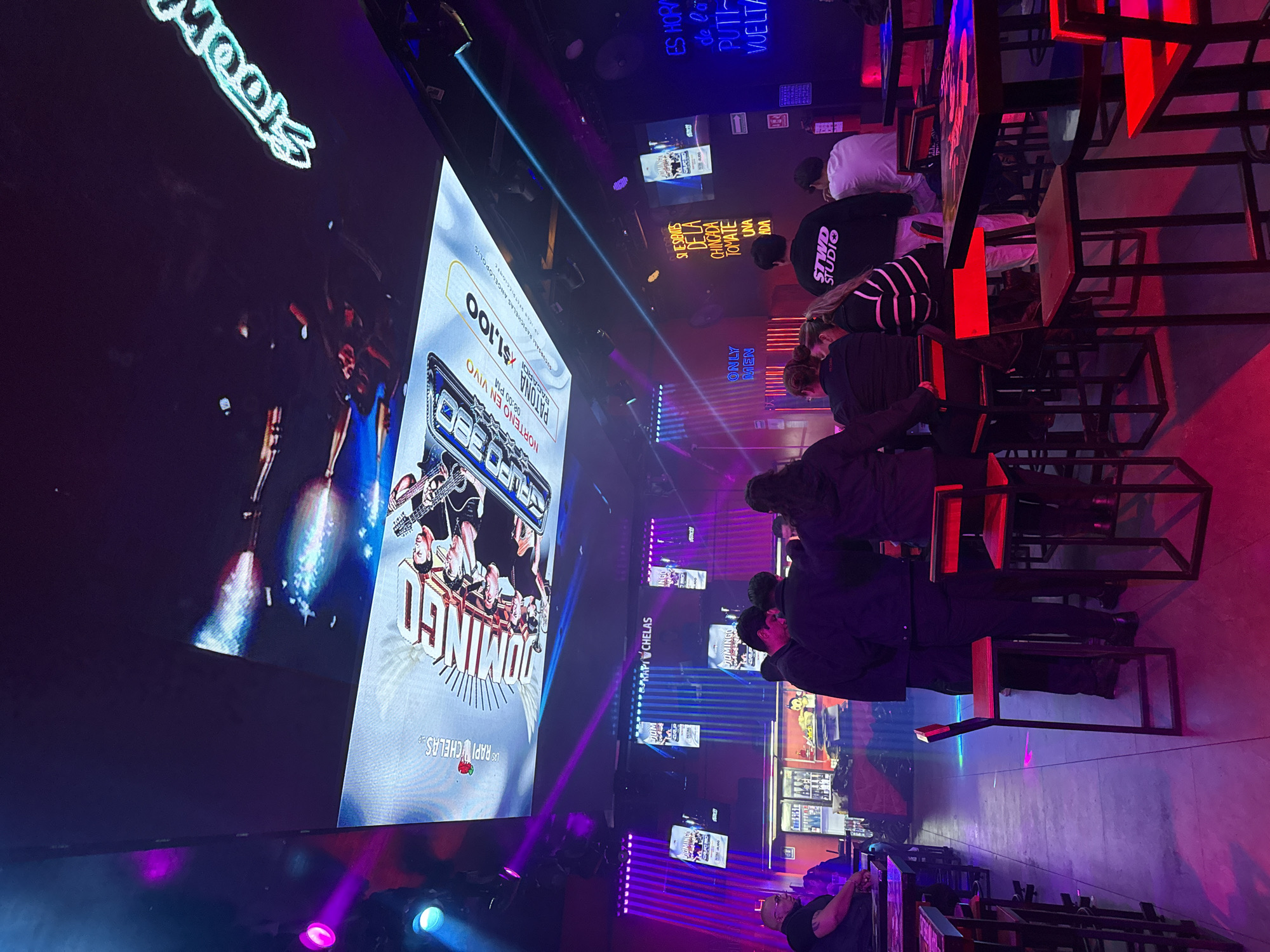 Here is a description of the image:
The image captures the inside of a crowded bar/restaurant, likely in Puebla, Mexico, based on textual cues. The scene is dimly lit, with neon signs and spotlights adding color and vibrancy. On the left, a large screen displays an advertisement for a band called "Grupo Domingo," scheduled to play "Norteno en Vivo" at 8 PM, with a price of 1,100 (presumably pesos). The location advertised is "Rapichelas Angelopolis." Smaller posters/advertisements are scattered on the walls behind the screen.
Several people are seated at tables and booths. The tables and chairs have a metal frame with wooden seats and tops. Customers, mostly facing the screen, are seated at the tables, their activities unclear. The overall atmosphere is lively and social, suggestive of an evening out.