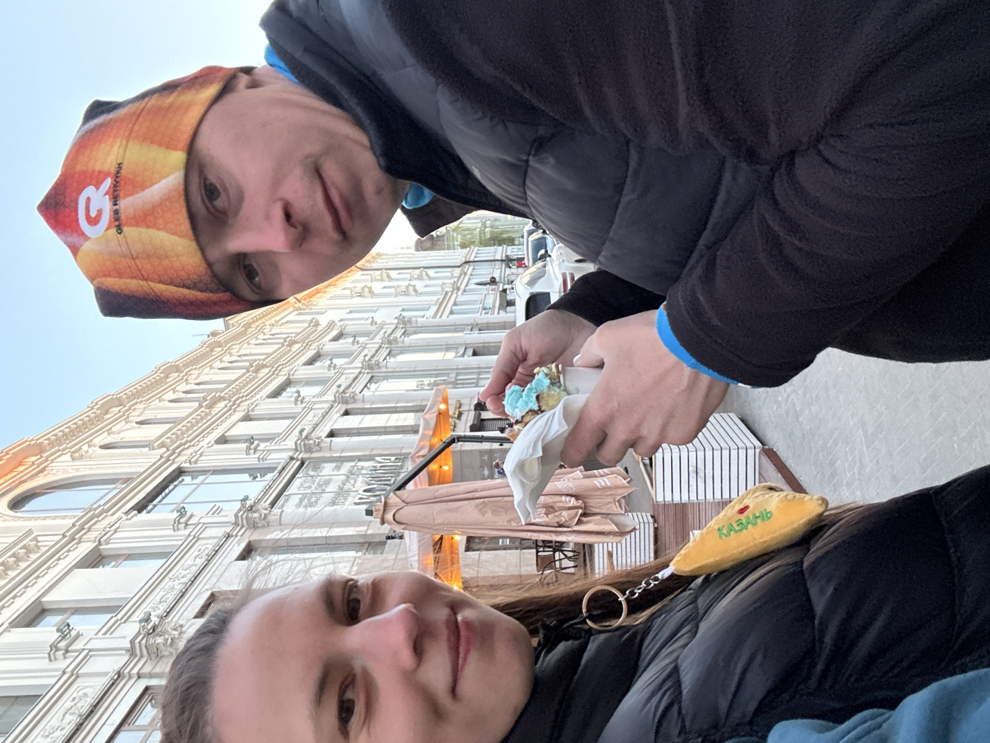 A selfie of two people, a male and a female, is taken in front of a building in Kazan, Russia. The female is in the foreground on the lower portion of the image, and the male is partially visible from above. Both individuals are wearing puffy black jackets. The male is also wearing a patterned beanie. He holds a snack wrapped in a white napkin. The building in the background is tall, with an ornate, light-colored facade and many windows. An awning and outdoor seating are partially visible below the building. A keychain with the word "Kazan" in Russian is attached to the female's jacket.