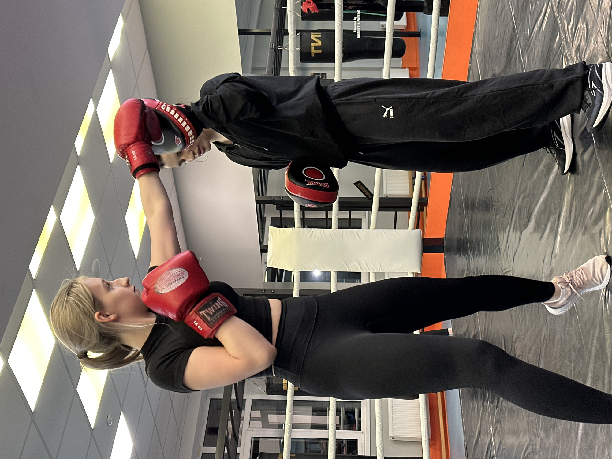 Two women are in a boxing ring. One woman, wearing a black top, black leggings, and pink sneakers, stands with her left arm raised, a red boxing glove covering her hand. She is looking at the other woman. The other woman, dressed in a black tracksuit with a Puma logo, faces the first woman. She has a red boxing glove on her left hand, which is touching the first woman’s glove. Her right hand holds a black and red striking pad. They are inside a boxing ring with white ropes, an orange frame, and a black floor. There are black punching bags hanging in the background, along with white and grey walls and windows. The location is a gym in the city of Kazan, Russia.