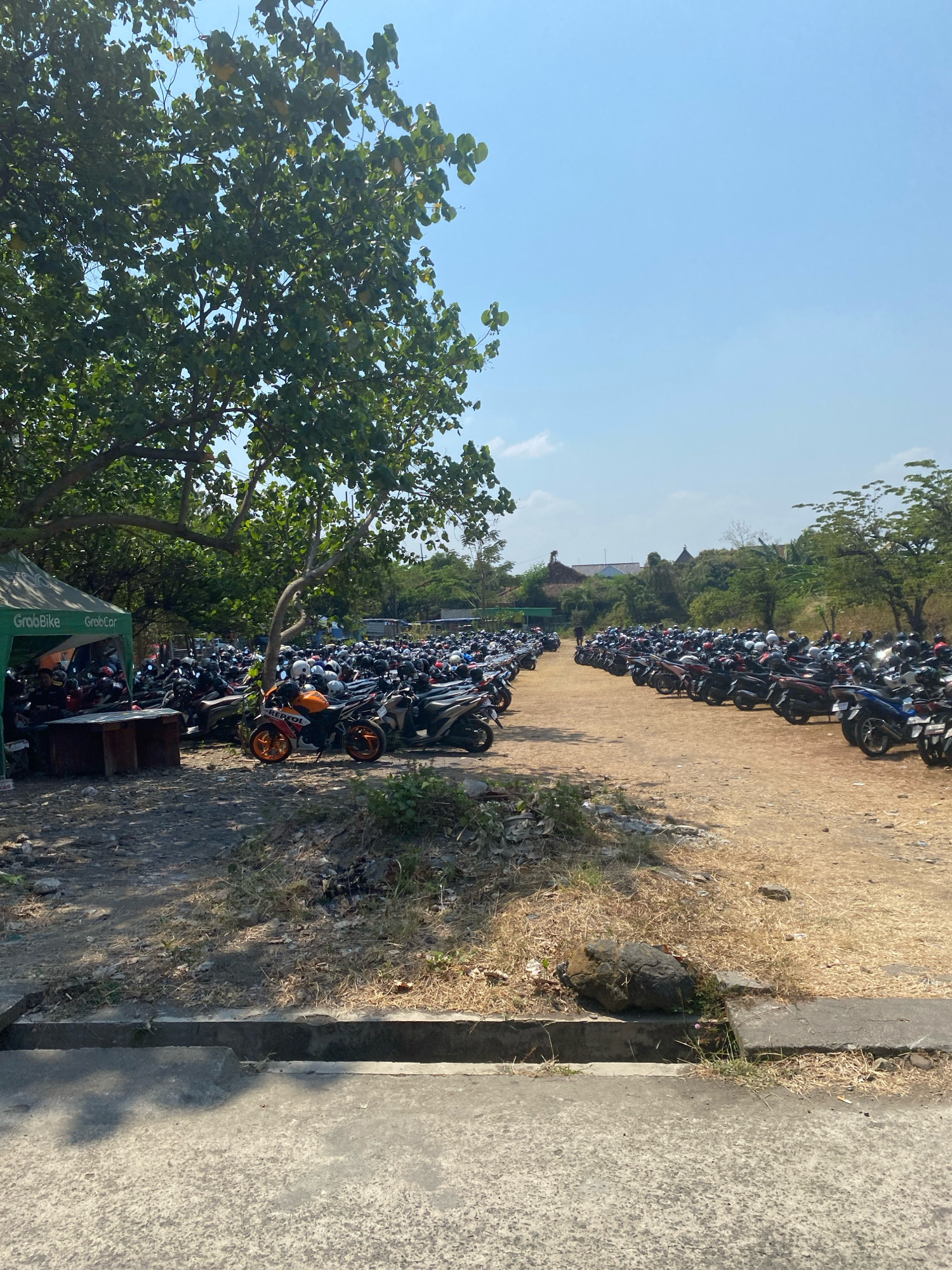 The image depicts an outdoor parking lot filled with a large number of motorcycles, likely in Kediri City, Indonesia. The lot is unpaved and dusty, with dry grass and some sparse vegetation. Several trees are present, casting shadows on the ground and some of the parked motorcycles.
On the left side of the image, a green tent with "GrabBike" and "GrabCar" branding is visible. Beneath the tent, there appears to be a table and some sort of structure, possibly a waiting area or service point. Several people are seen near the tent and within the parking lot, though their specific actions are not discernible due to the distance and resolution.
The motorcycles are parked in rows, occupying a significant portion of the frame. They are varied in color and model, with many of them appearing to be scooters. Helmets are visible on some of the handlebars. The sky is clear and blue, indicating a sunny day. In the background, there are some buildings and more trees, suggesting a somewhat developed area. The foreground shows a concrete path or road with a drainage ditch.