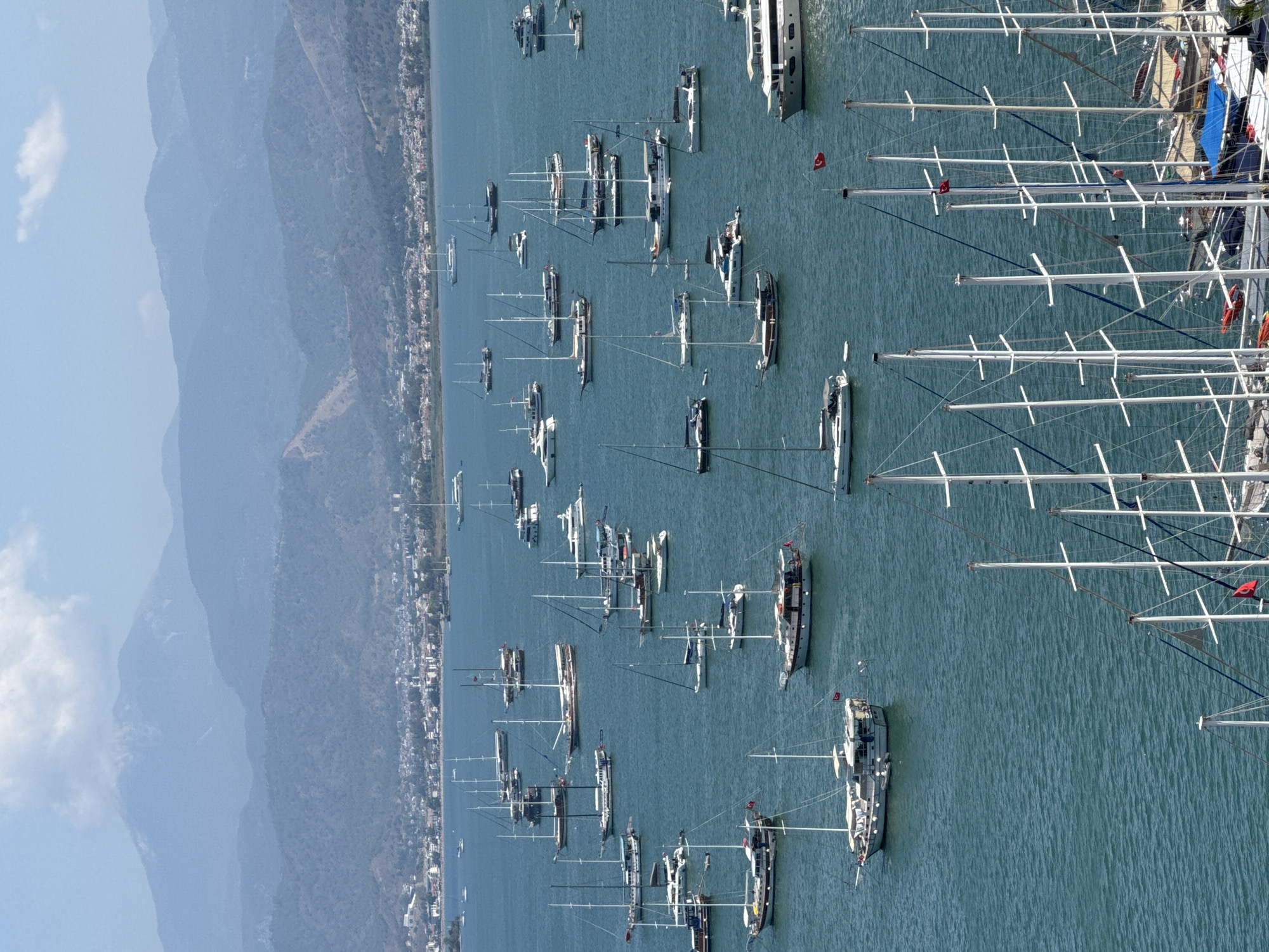 This image captures a wide-angle, elevated view of a bay in Fethiye, Turkey, under clear daylight conditions. The scene is dominated by a vast expanse of blue-green water filled with numerous boats, with a coastal city extending along the far shoreline and multiple mountain ranges receding into the distant background.
In the immediate foreground, the white masts and intricate rigging of several large sailing vessels are partially visible, suggesting a proximity to a viewing point or a densely packed mooring area.
The mid-ground features a calm body of water teeming with a diverse fleet of vessels. Over a hundred boats are present, predominantly consisting of sailing yachts (including traditional wooden gulets and modern sloops) and several multi-deck motor yachts. Many boats are white-hulled, some featuring visible cabins and deck awnings. Multiple Turkish flags (red with a white star and crescent) are visible flying from the masts or sterns of various boats. The boats appear to be stationary, either at anchor or moored, distributed across the bay without discernible patterns of movement.
Along the curved distant shore, a sprawling coastal city is evident, characterized by numerous light-colored buildings with scattered red-tiled roofs, nestled against verdant, undulating hills. The urban sprawl extends along the waterfront and up the lower slopes of the hills.
In the background, a series of progressively lighter blue-grey mountain ranges rise, creating an effect of atmospheric perspective. The nearest mountains are detailed with forested slopes, while the farthest peaks are hazy and indistinct, with a few faint white clouds clinging to the highest elevations. The sky above is a bright, clear blue, with a few sparse white cumulus clouds. No individual people or specific actions are discernible at this resolution.